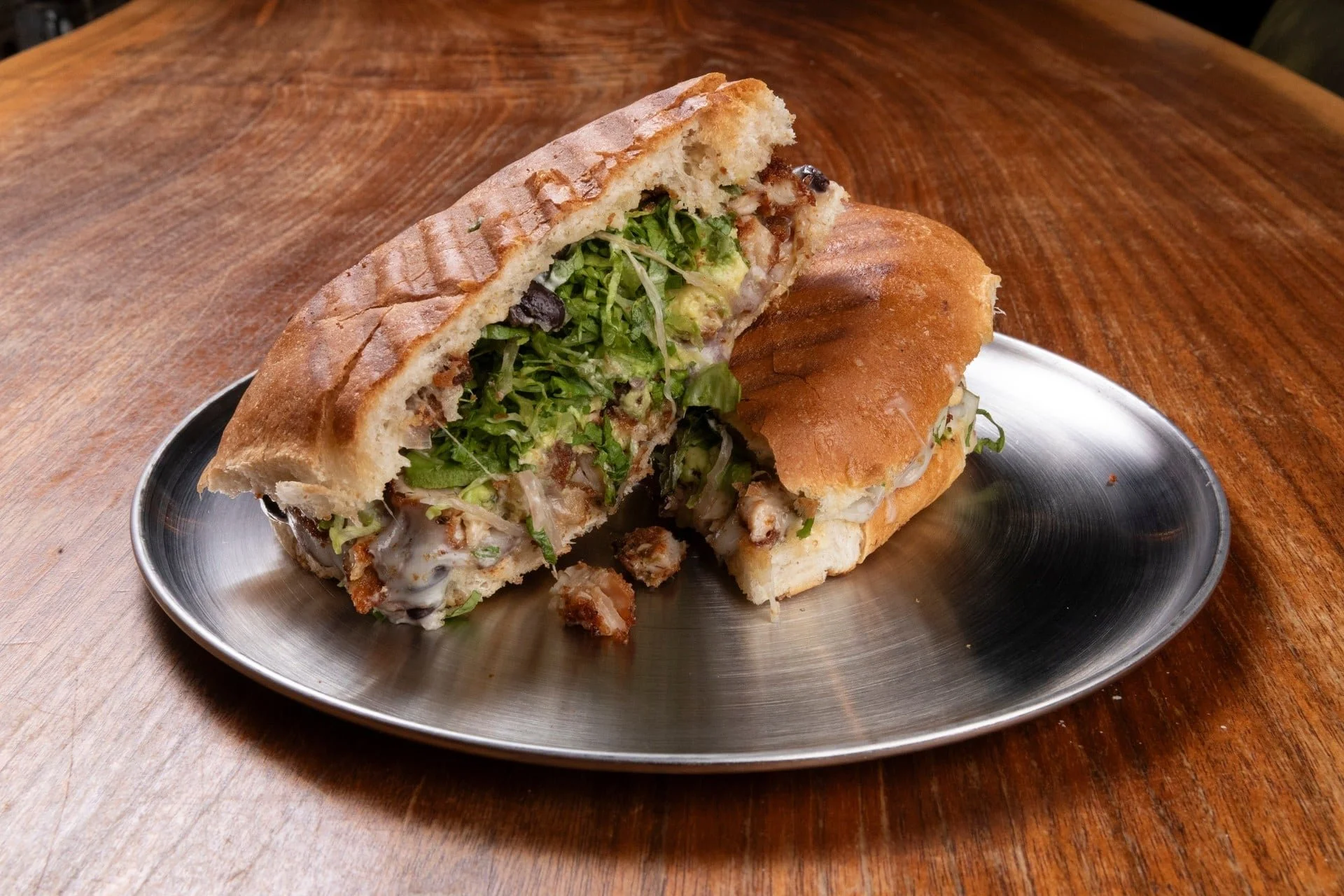 A large sandwich or sub on a metal plate with lettuce, other greens, and possibly meats and cheese inside, on a wooden table.