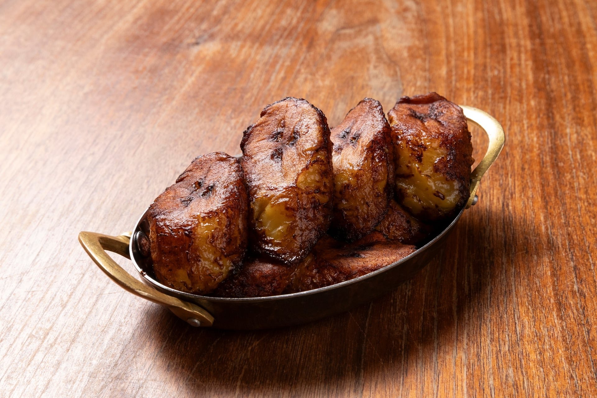 Four pieces of grilled plantains with caramelized edges served in a silver dish on a wooden surface.