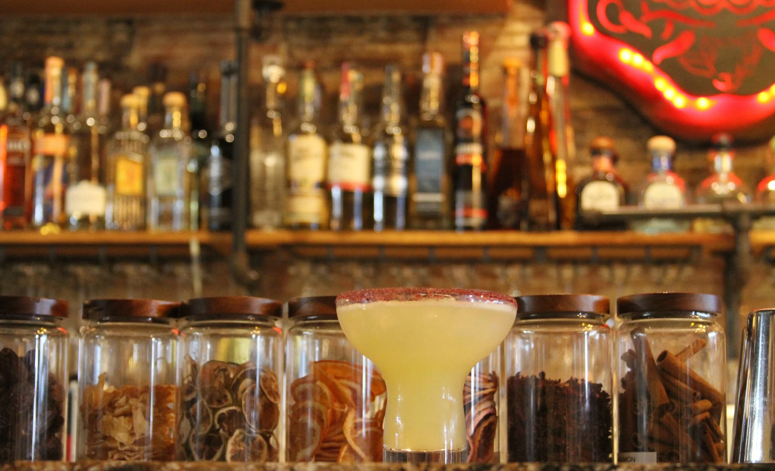 A yellow cocktail drink in a coupe glass with red seasoning on the rim, placed on a bar counter with shot glasses filled with various ingredients behind it, and a blurry background of liquor bottles on a wooden shelf.