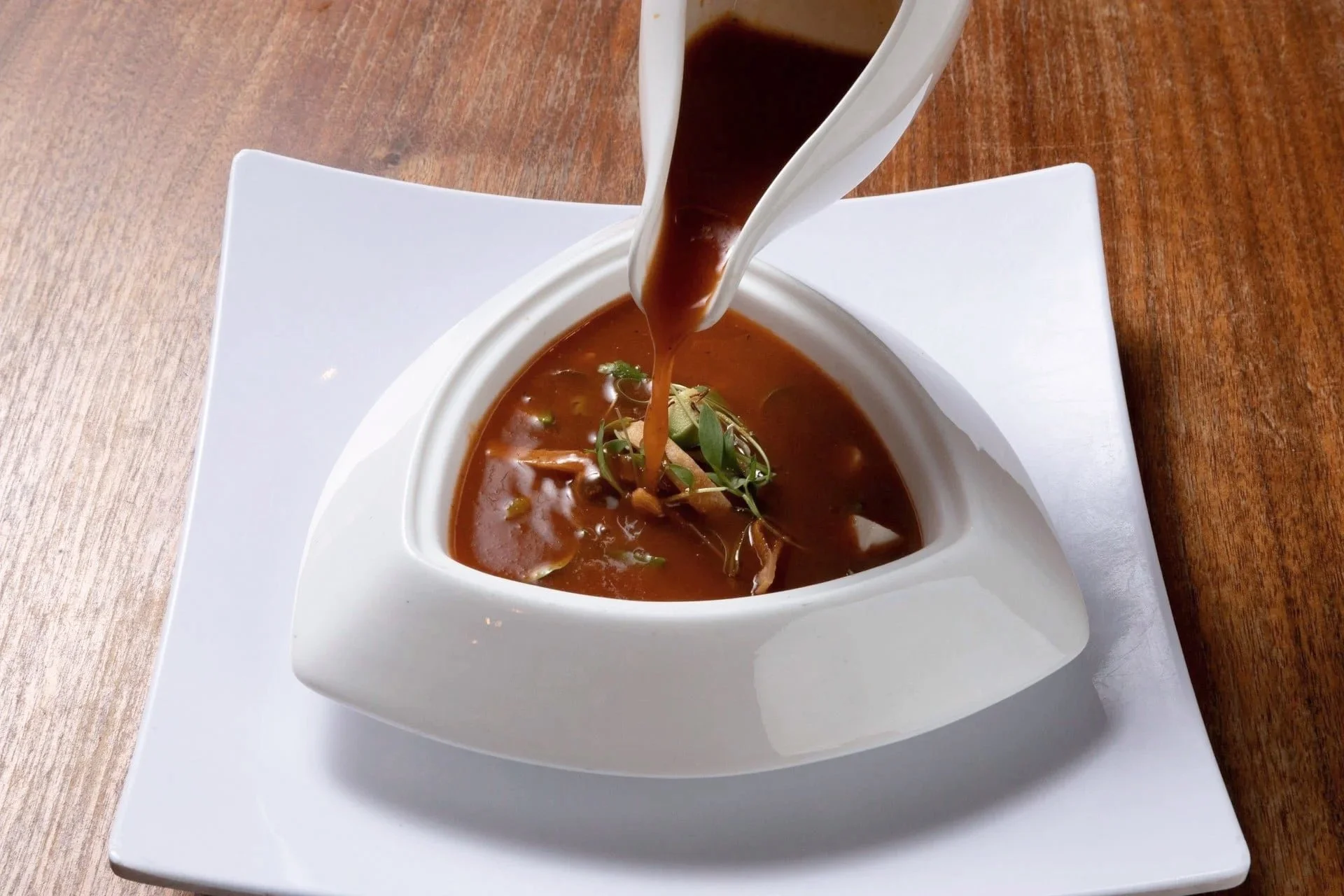 A bowl of dark brown soup garnished with green herbs, served in a white bowl on a square white plate on a wooden table.