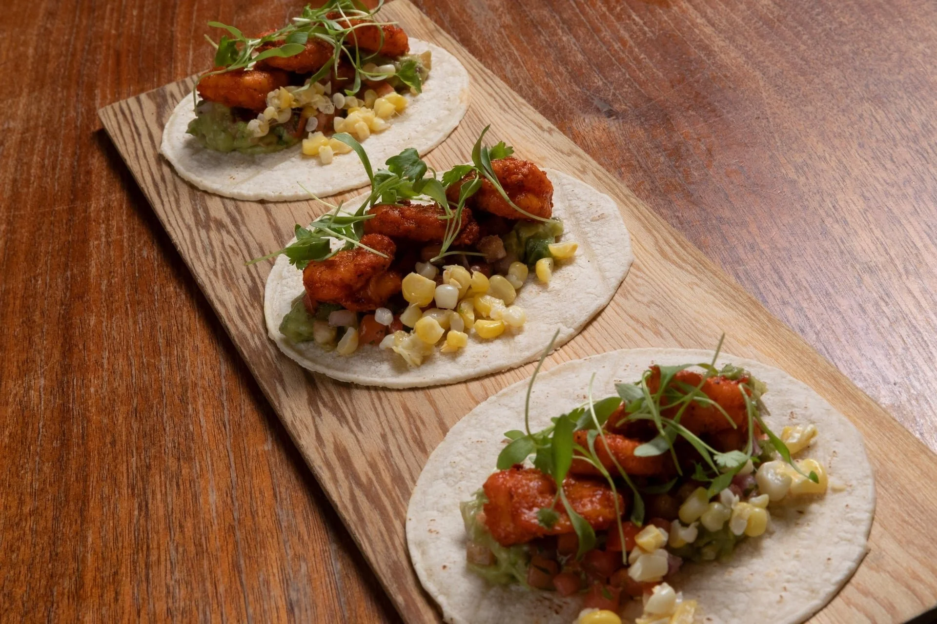 Close-up of three tacos on a wooden serving board, filled with fried chicken, corn, chopped vegetables, and microgreens, on a wooden table.