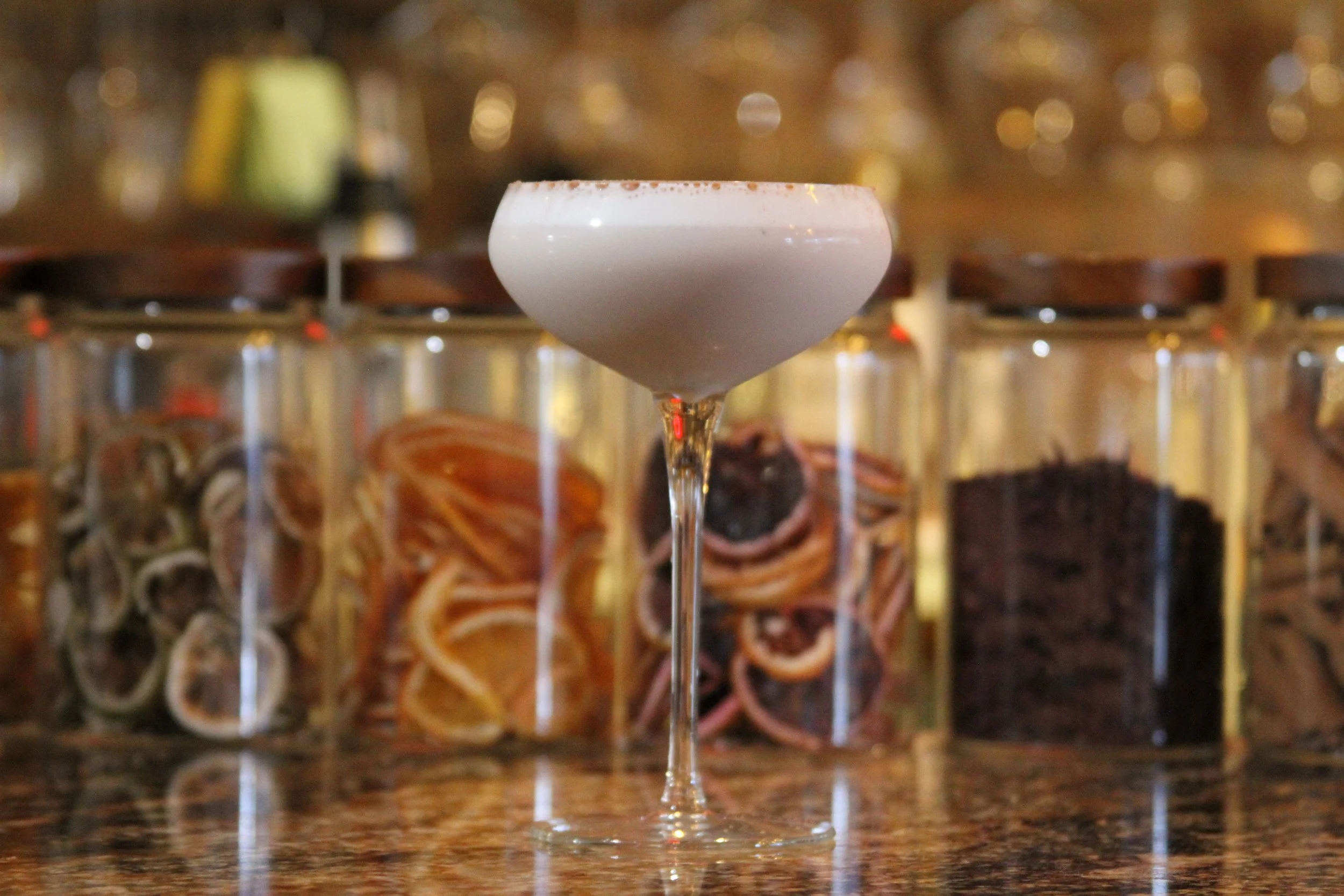 A white cocktail in a coupe glass with a salted rim on a bar counter, with blurred bottles in the background.