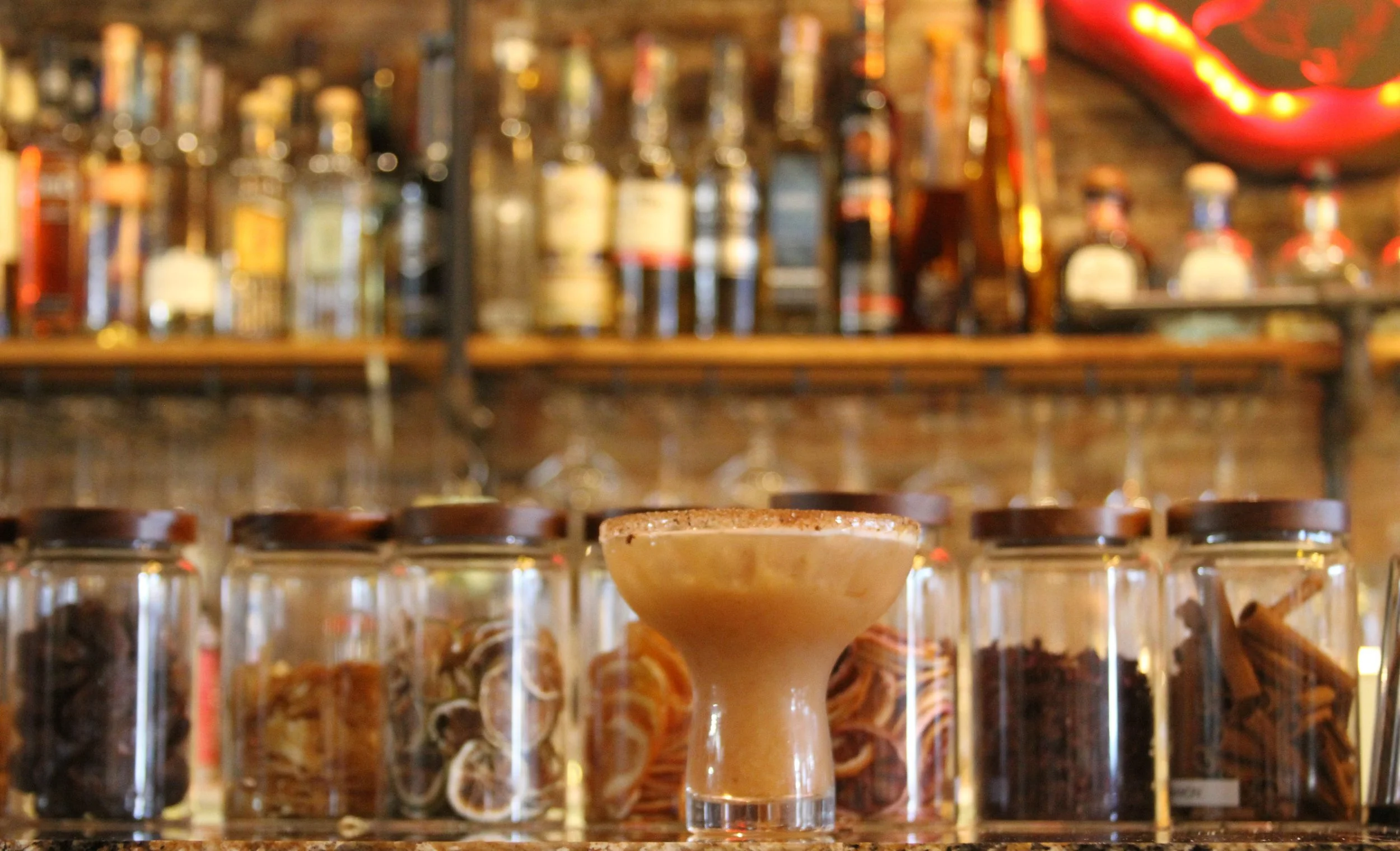 Blurred background of liquor bottles on a bar shelf with a cream-colored margarita glass in the foreground.