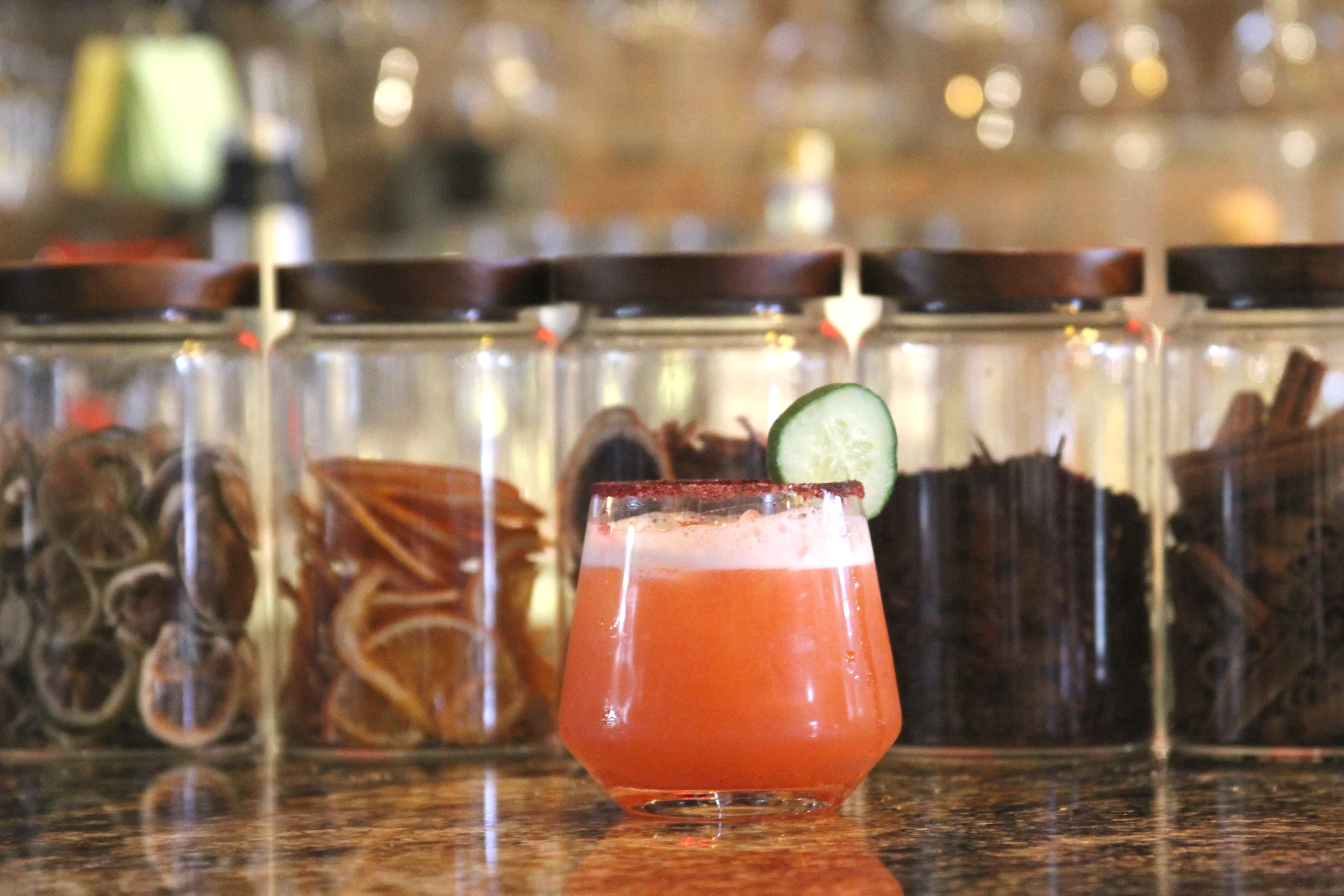 Close-up of a pink cocktail garnished with a slice of cucumber, served in a rounded glass on a granite countertop with a blurry bar background.