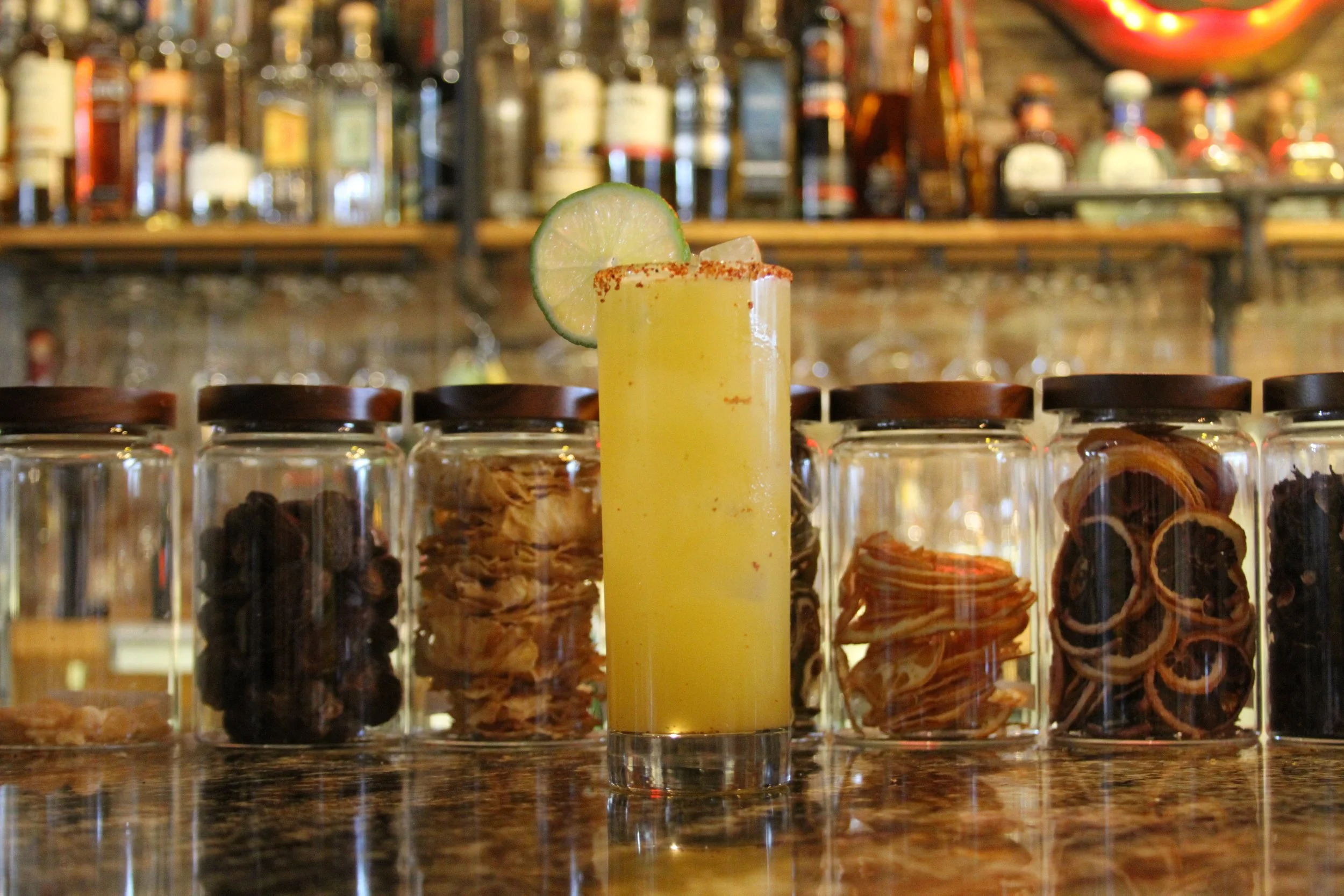A yellow cocktail garnished with a lime wheel and red chili powder rim, set on a bar counter with glass jars and a blurred bar background.