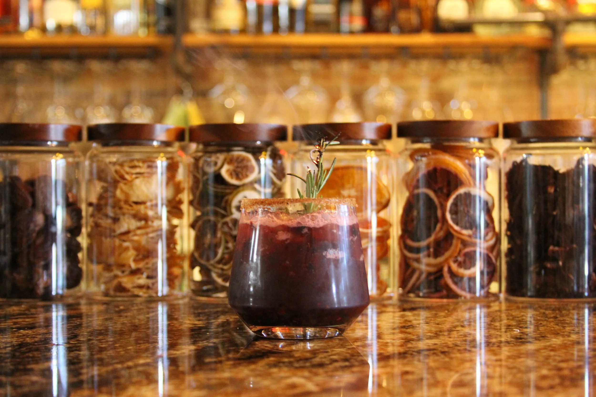 A small glass filled with a dark red berry-based beverage, garnished with a sprig of rosemary, placed on a polished wooden surface. Behind it, there are several glass jars filled with dried herbs or fruit, and a blurred background of bottles on wooden shelves in a cozy bar or kitchen setting.
