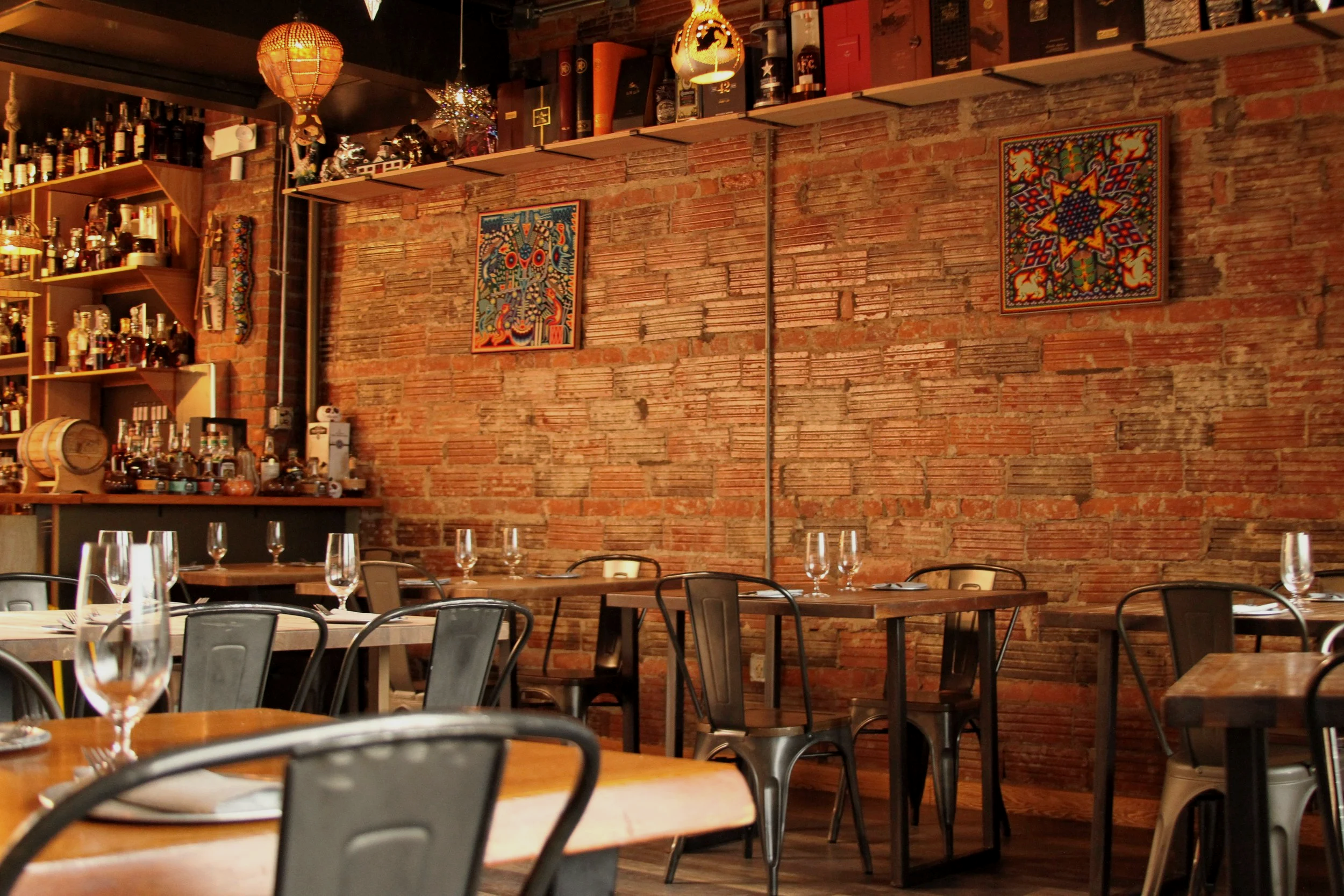 A cozy restaurant with a brick wall decorated with colorful artwork and shelves of liquor bottles. Wooden tables are set with glasses, and black chairs are arranged around them. Warm lighting creates an inviting atmosphere.