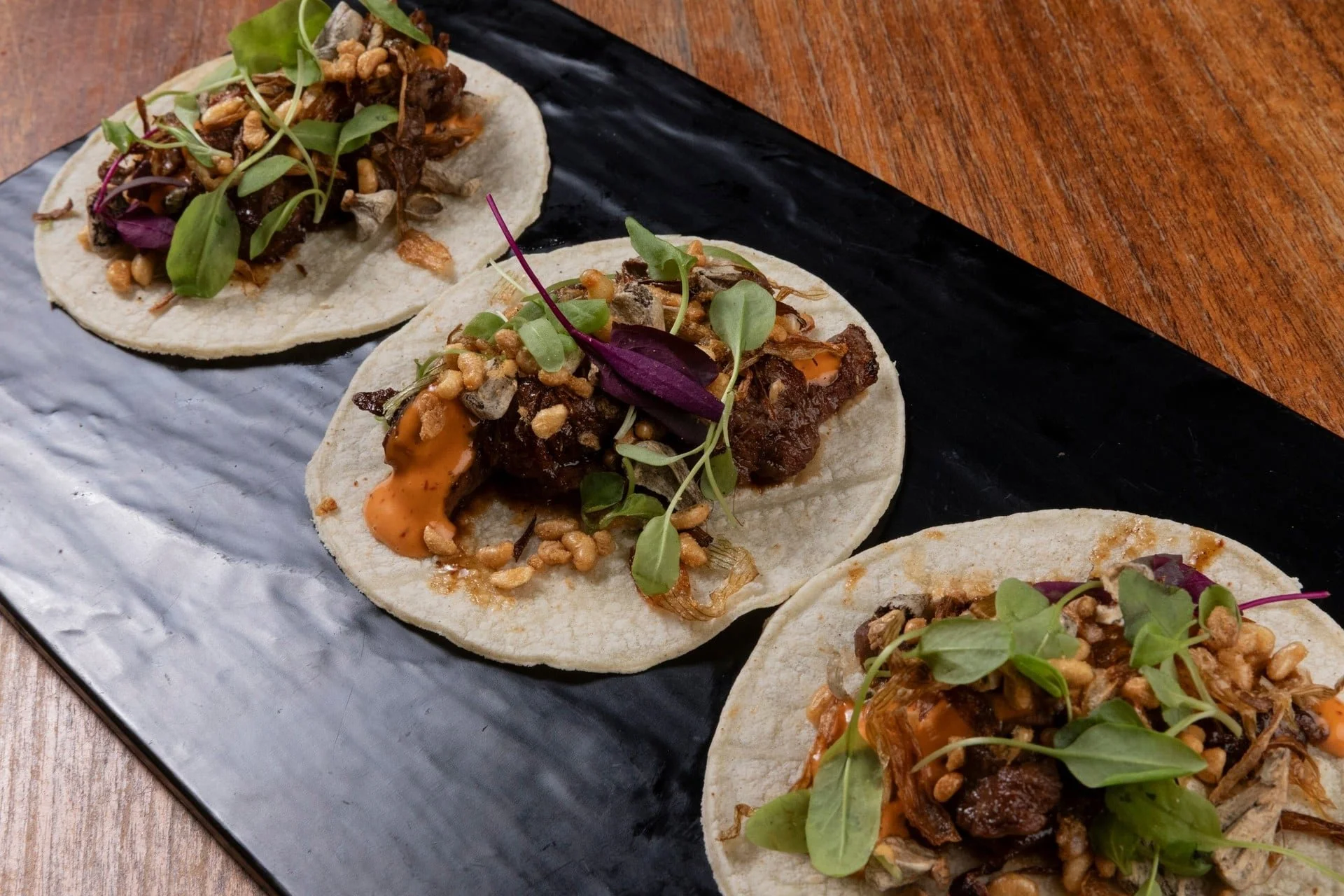 Close-up of three small tacos with beef, microgreens, and crunchy toppings on a black rectangular plate on a wooden table.