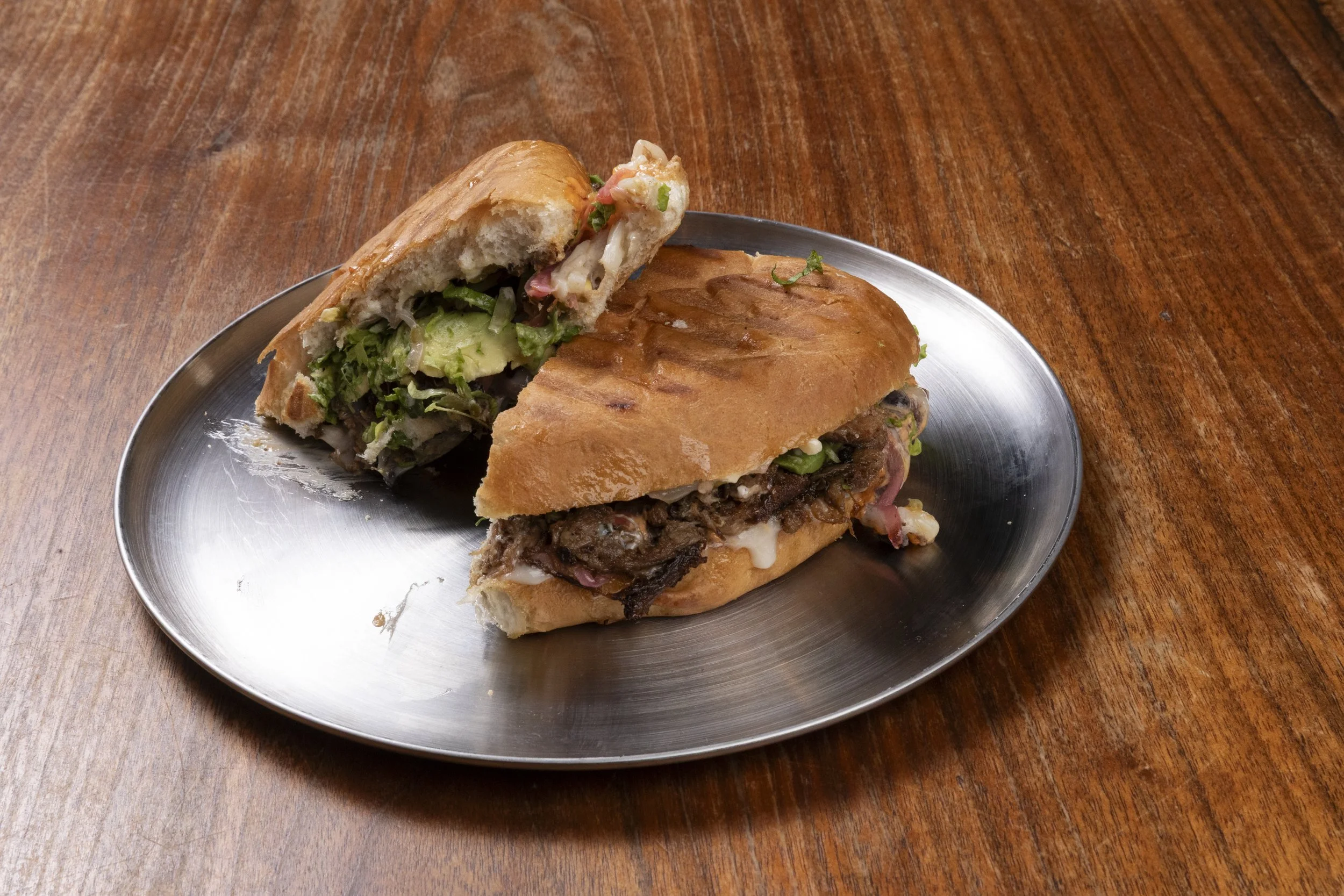 A metal plate with two sandwiches on a wooden table. One sandwich is cut in half, showing lettuce, tomato, and meat inside a baguette. The other sandwich is whole, with visible meat and vegetables.