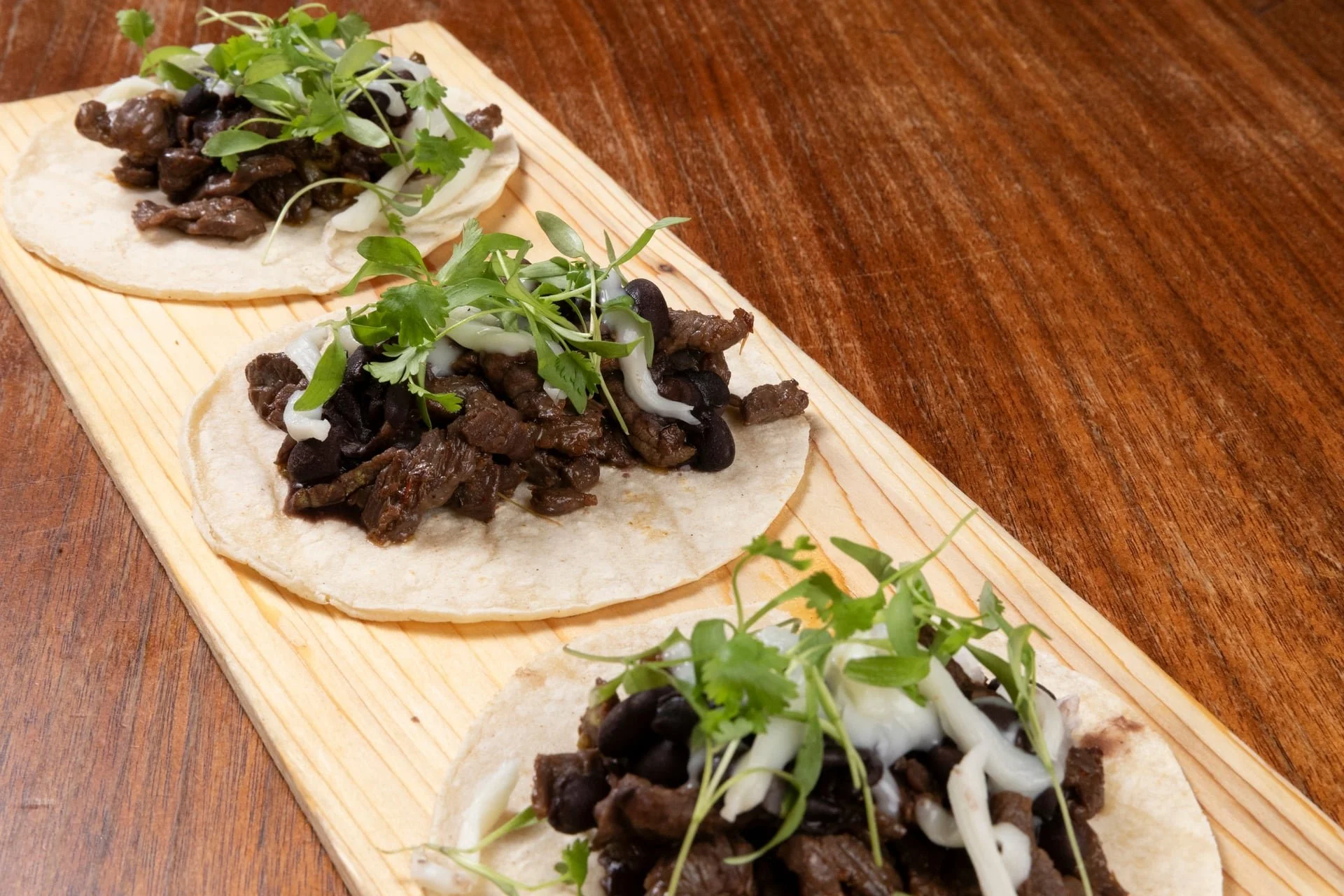 Three street tacos topped with green herbs on a wooden serving board.