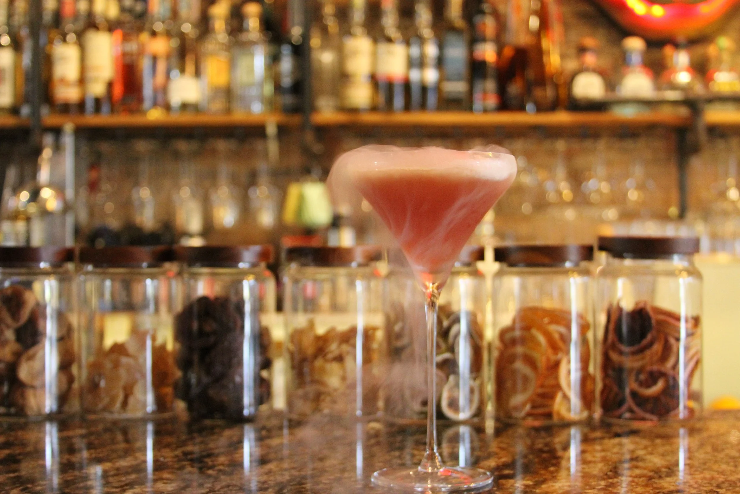 A pink frozen cocktail in a martini glass on a bar counter, with blurred bottles of alcohol in the background.