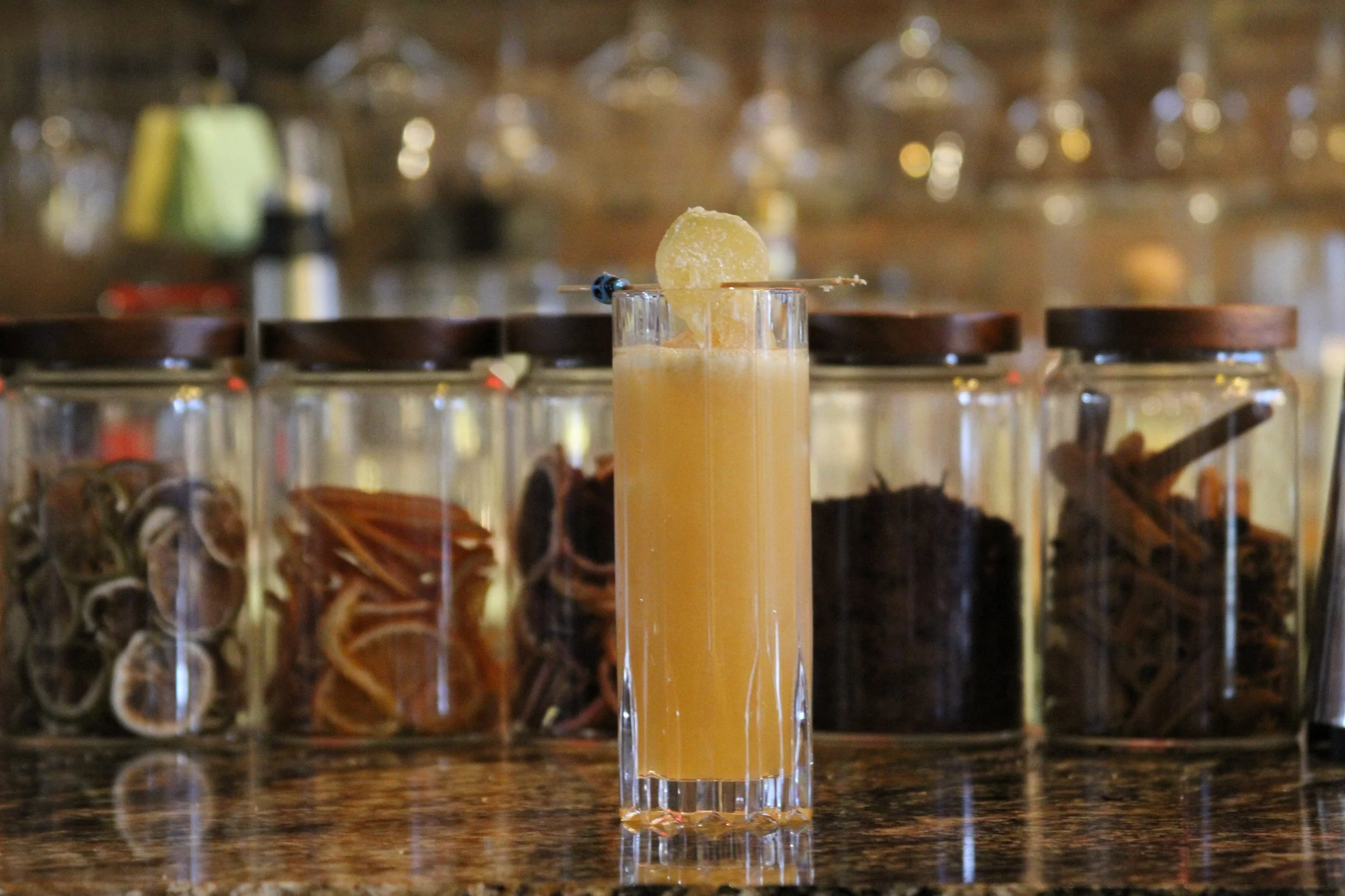 A tall glass of orange-colored beverage with a lemon slice garnishing the rim, placed on a kitchen or bar counter with a blurred background of glass jars and bottles.