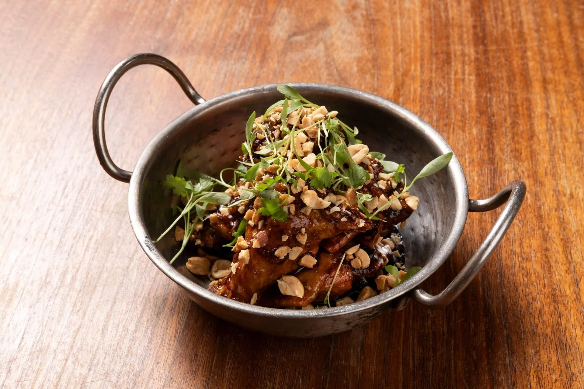 A silver oval serving dish with handles holding a glazed meat dish topped with chopped nuts and garnished with microgreens, placed on a wooden table.