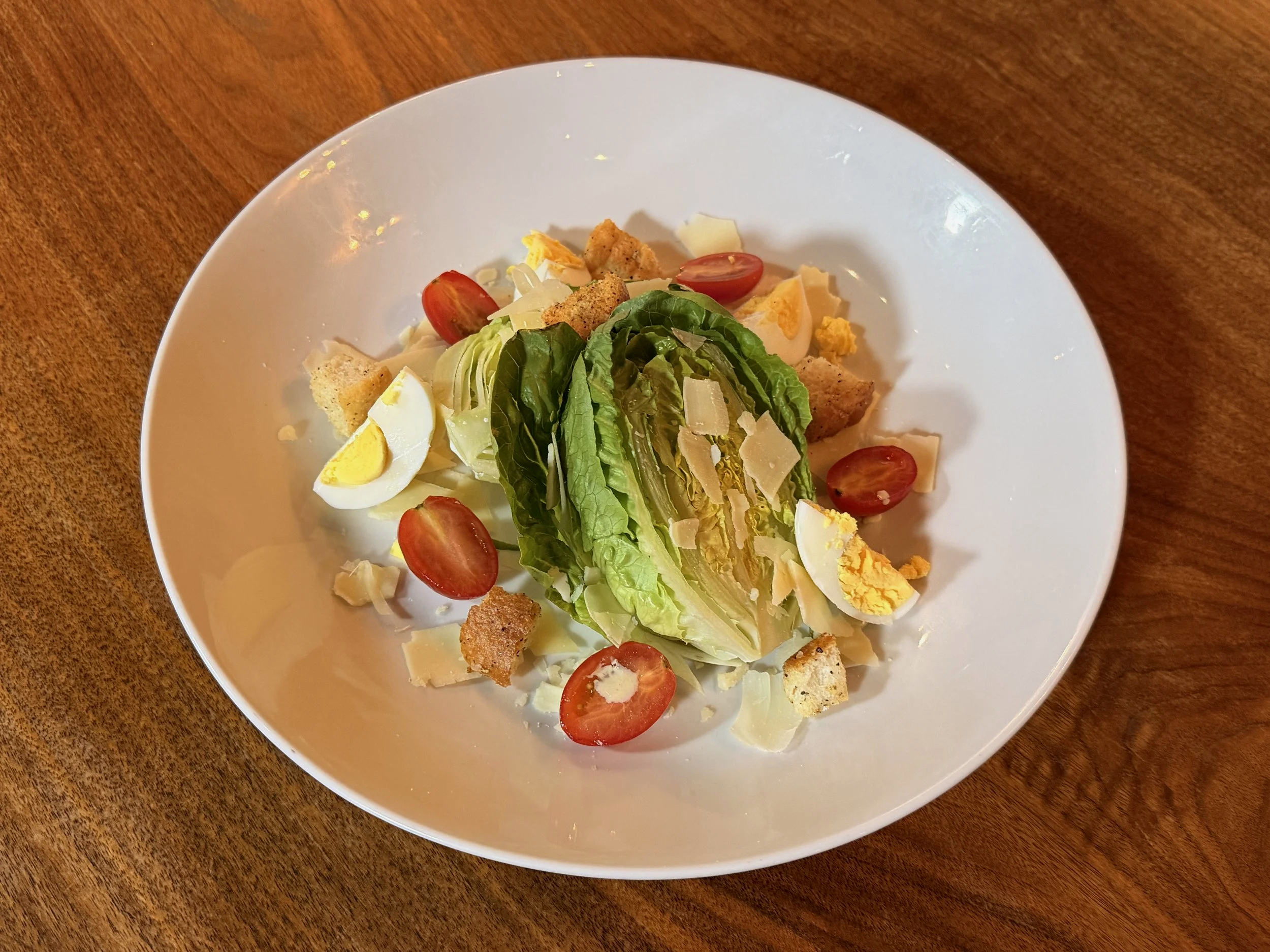 A white bowl containing a salad with cherry tomatoes, sliced boiled eggs, lettuce, shredded cheese, and croutons on a wooden table.