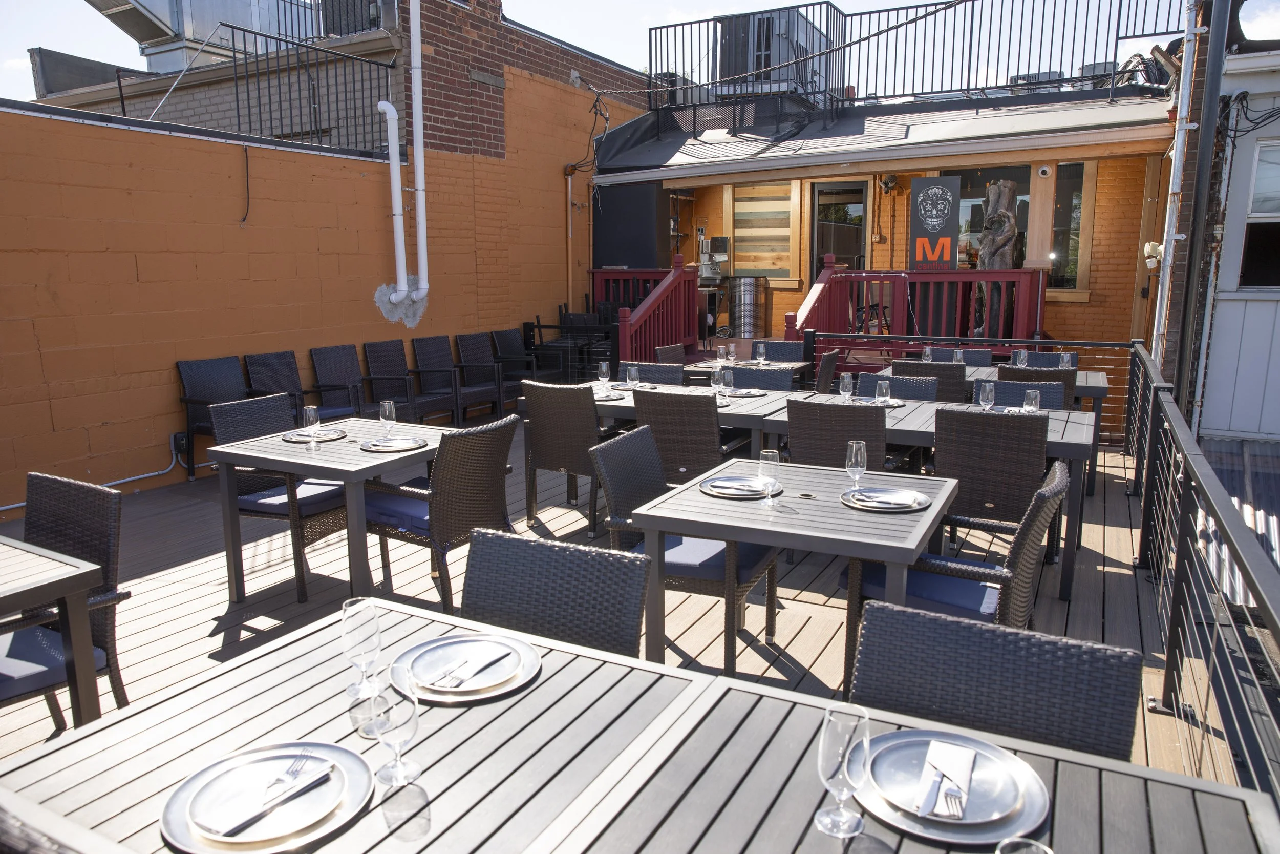 Outdoor rooftop patio with tables set for dining, including plates and glasses. There is a brick wall painted orange and some chairs stacked against it. The patio is enclosed by metal railings and has a small raised area with stairs, and decorative elements including a large art piece and a wooden sculpture.