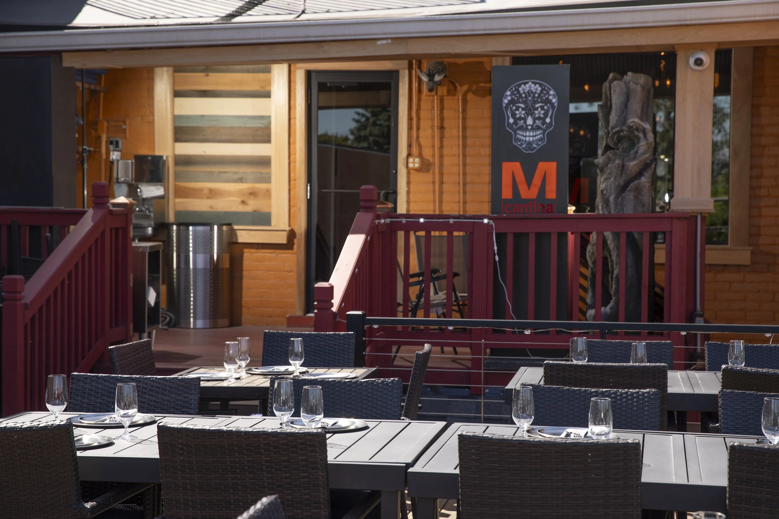 Outdoor patio of a restaurant or bar with tables and chairs set with wine glasses. There's a wooden railing and a building with brick and wood accents, and a sign with a skull design and the word 'cantina' on it.