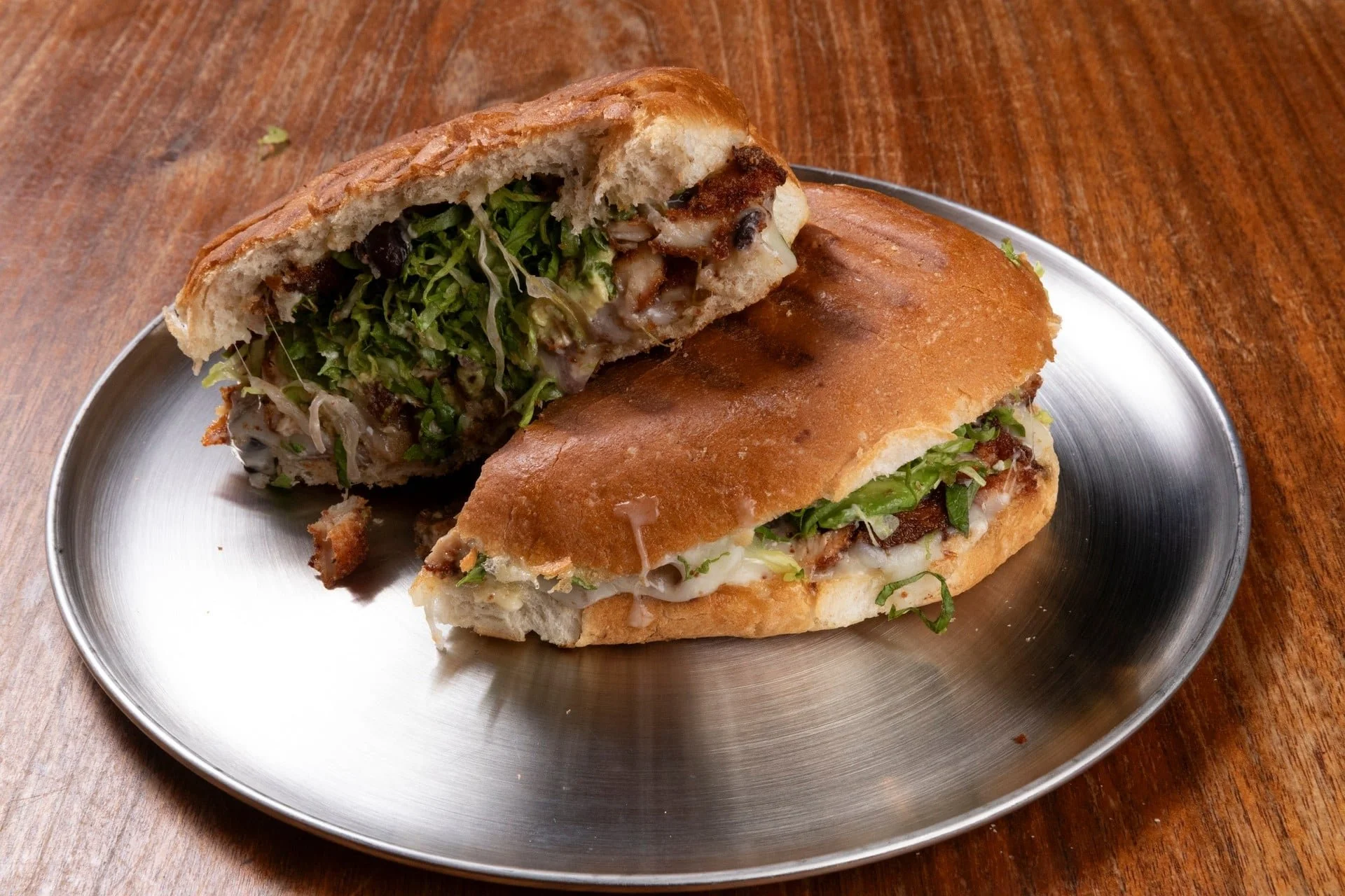 Half of a sandwich with lettuce, cheese, and meat on a bun, served on a metal plate on a wooden table.
