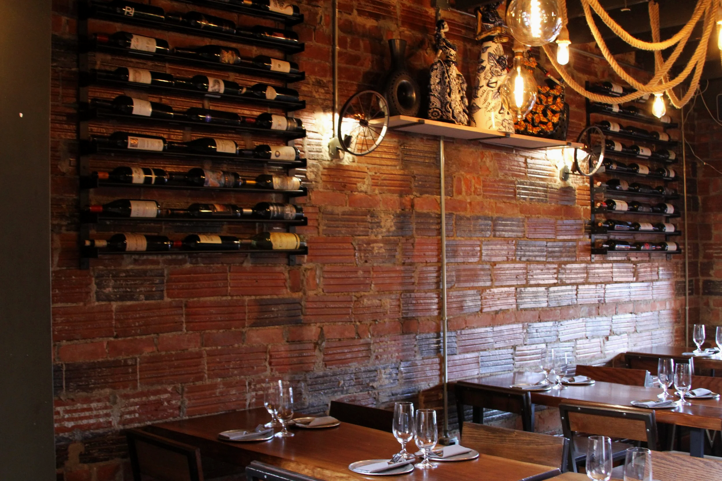A cozy restaurant interior with a red brick wall decorated with black metal wine racks holding several bottles of wine. The wall also features rustic shelves displaying fabric-covered objects and vintage metal wheels. Warm lighting from hanging light bulbs adds to the inviting atmosphere. Wooden dining tables are set with wine glasses, plates, and silverware, with chairs arranged around them.