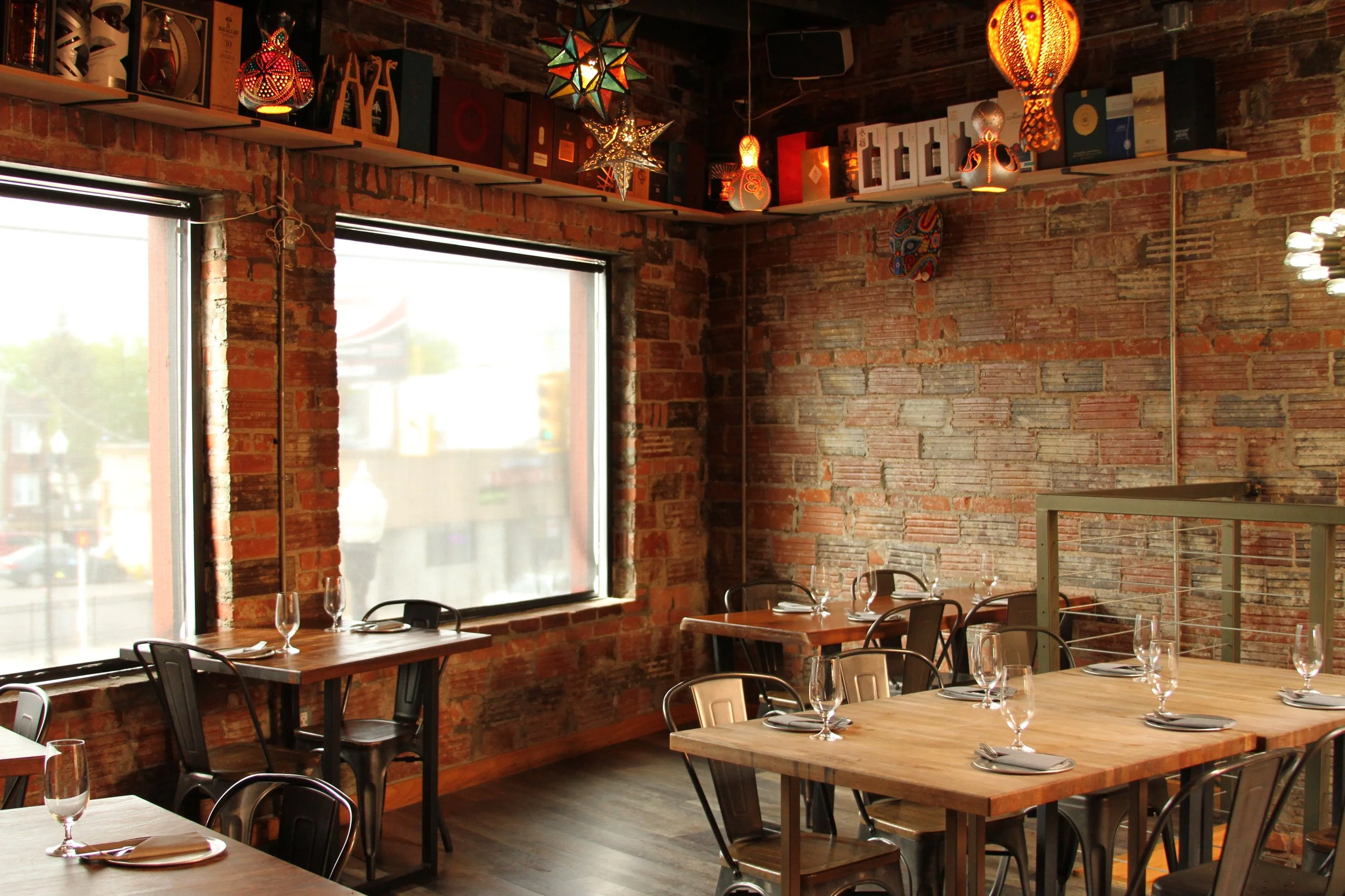 Interior of a restaurant with brick walls, large window, and hanging colorful star-shaped lanterns and lamps, set with tables and wine glasses.