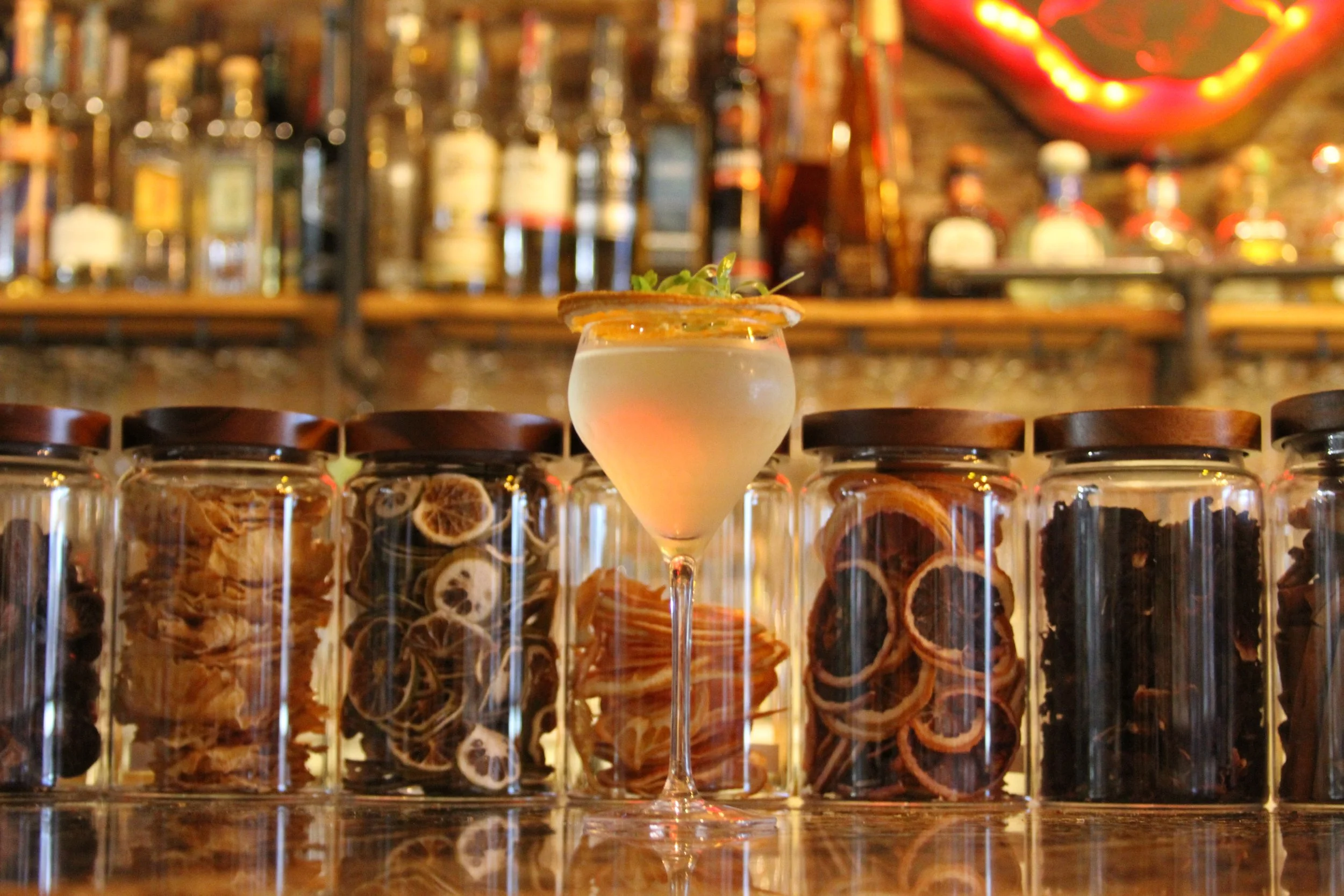 A cocktail in a martini glass filled with a white beverage, garnished with pink and orange slices and a small green herb on top. The drink is positioned in front of a row of glass jars filled with dried herbs or fruits, and the background shows a bar with bottles and colorful decorations.