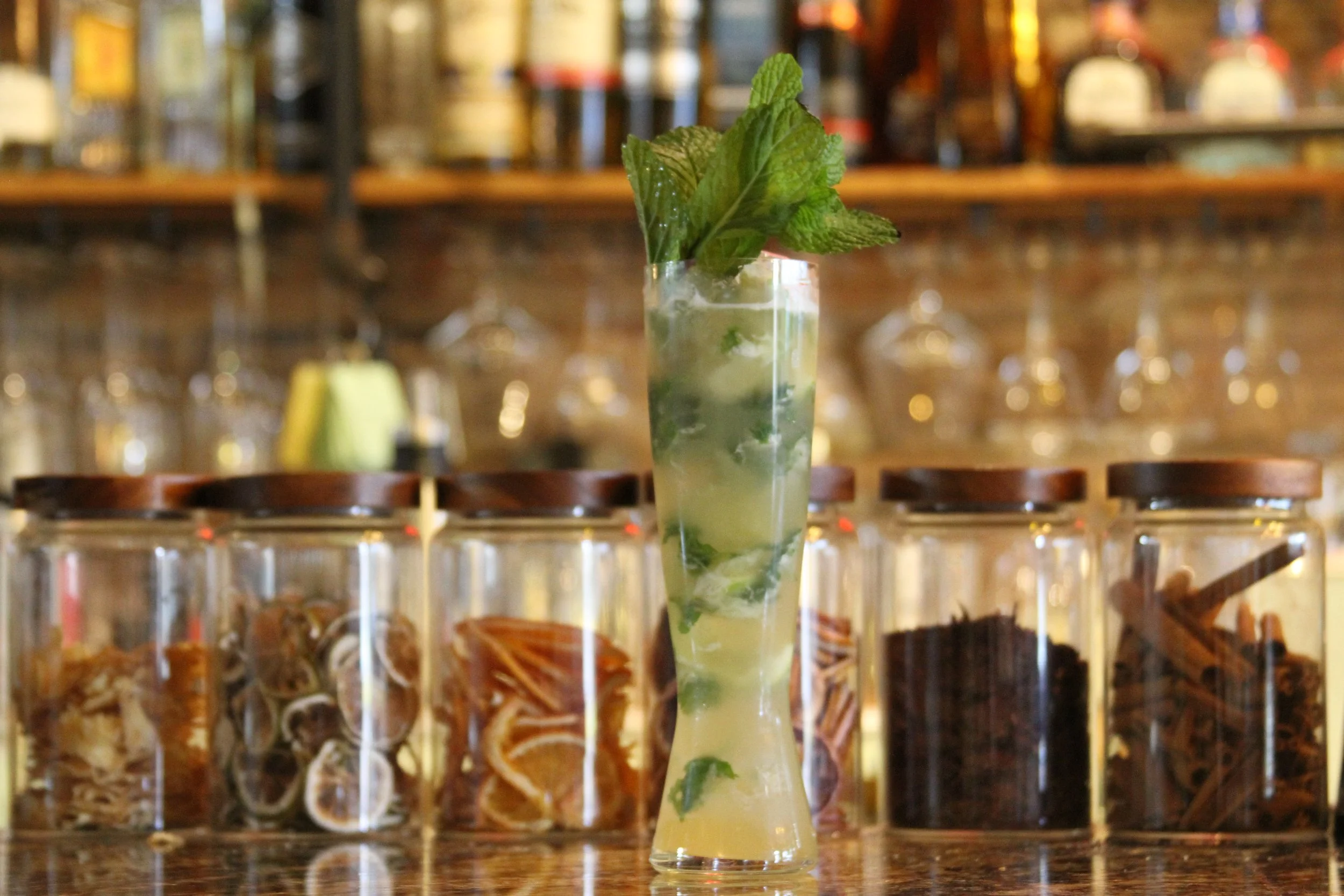A tall glass of mojito with mint leaves on top, set on a bar counter with blurred liquor bottles in the background.