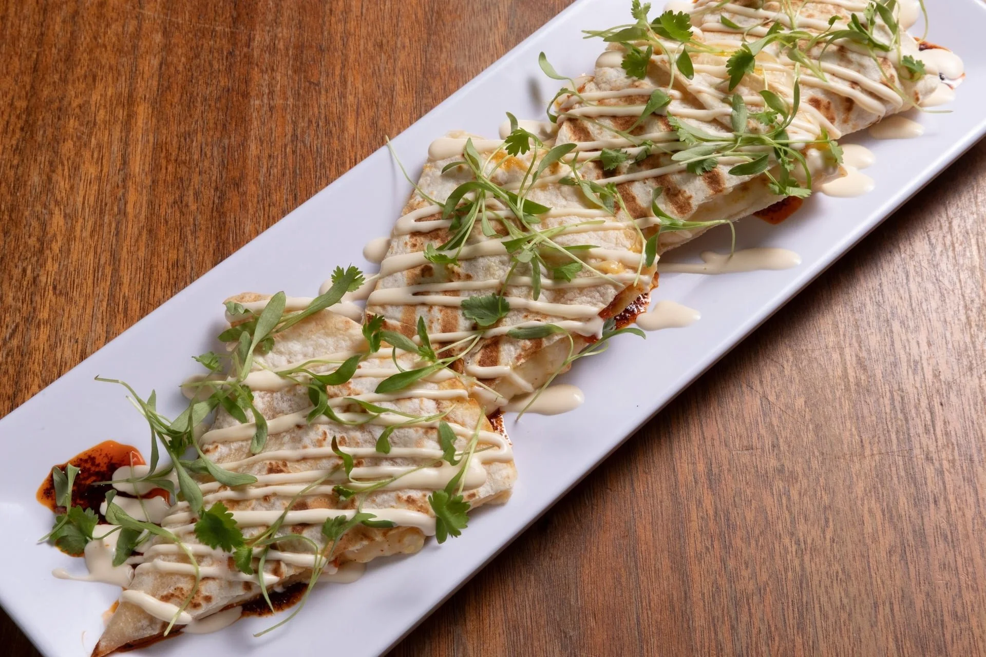 A rectangular white plate of grilled chicken wraps garnished with microgreens and drizzled with creamy sauce and hot sauce, placed on a wooden table.