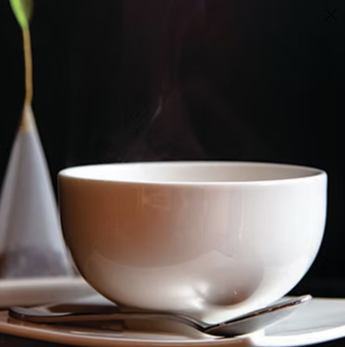 A steaming white bowl on a plate with a spoon next to it and a teapot in the background