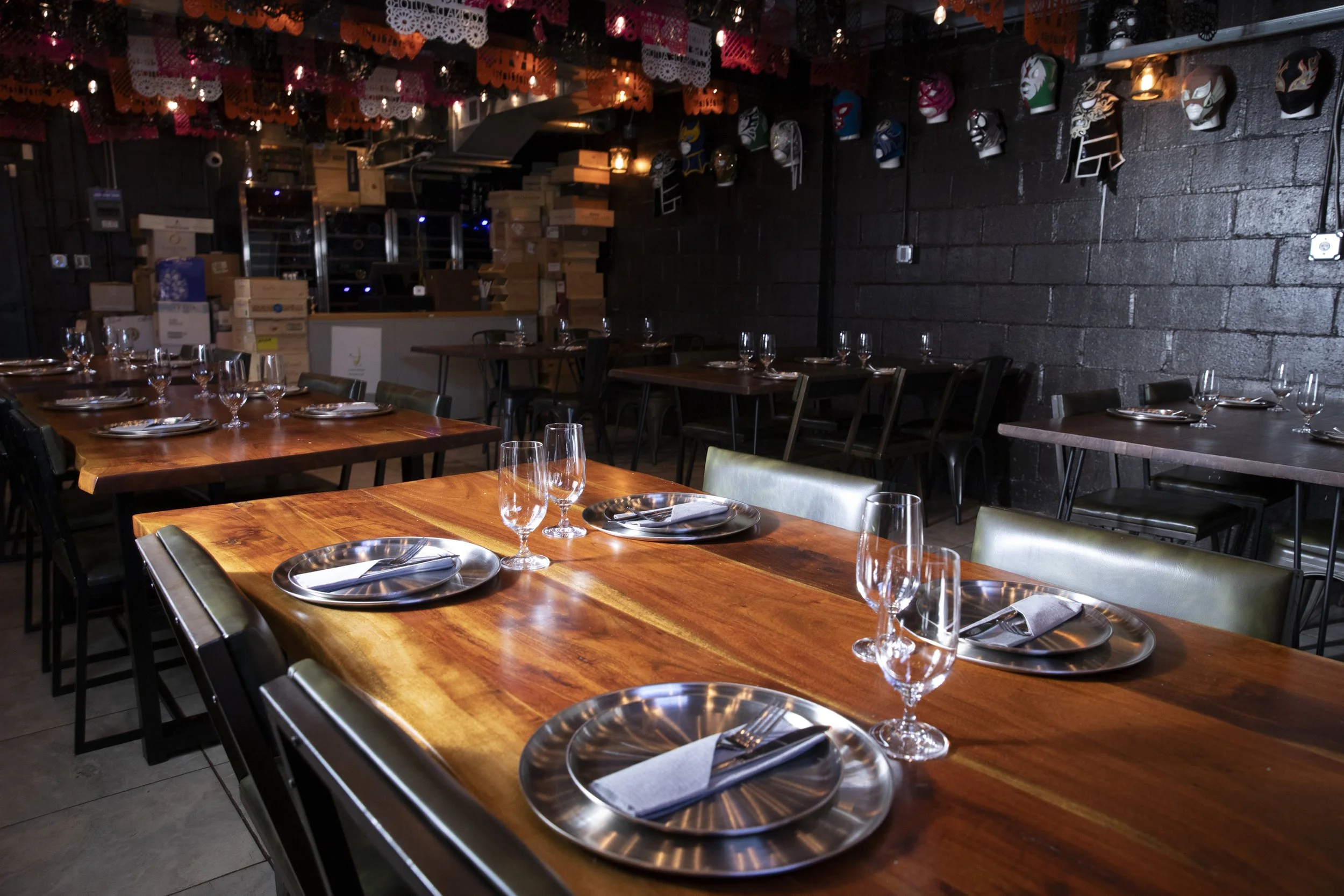 Interior of a restaurant with wooden tables set with plates, forks, knives, wine glasses, and napkins. Decorated with colorful papel picado banners and masks on the black brick wall.