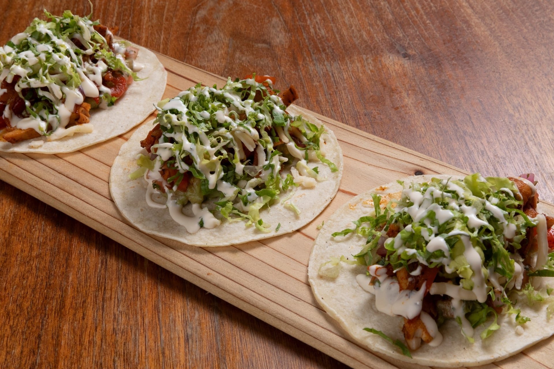 Three tacos with shredded lettuce, grilled chicken, tomatoes, cheese, and drizzled with white sauce on a wooden serving board.