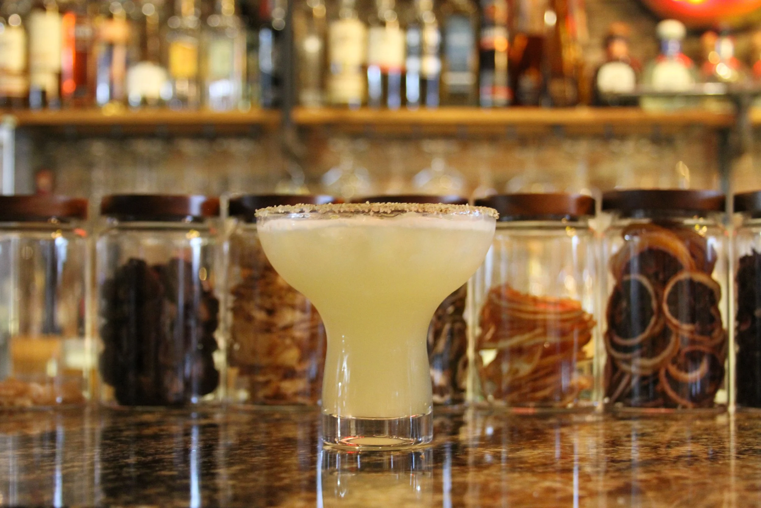 A creamy yellow cocktail garnished with a salted rim, set on a bar counter with a blurred background of liquor bottles and bar shelves.