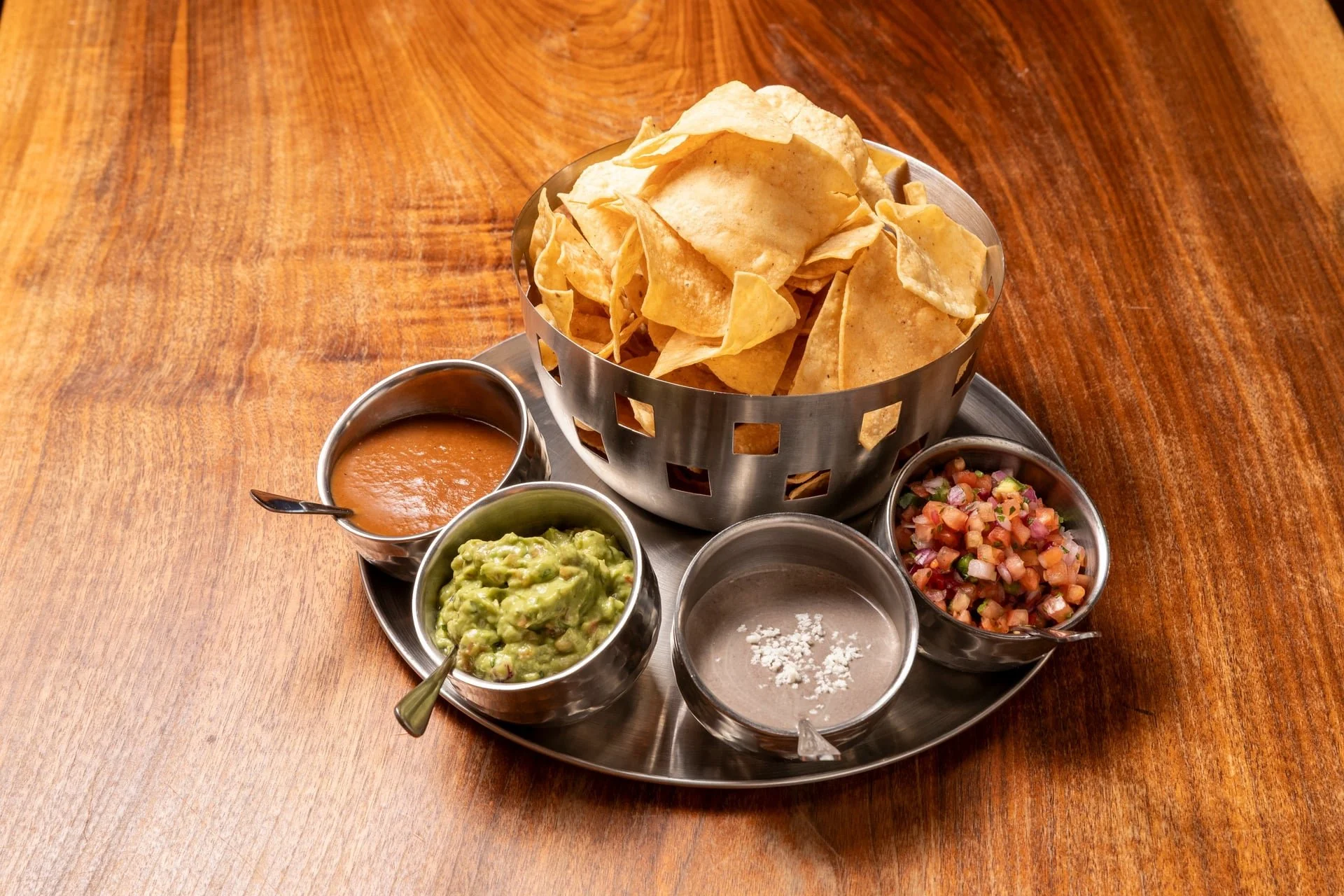 A metal tray with tortilla chips, guacamole, salsa, and pico de gallo, along with small metal cups of sauce, on a wooden table.