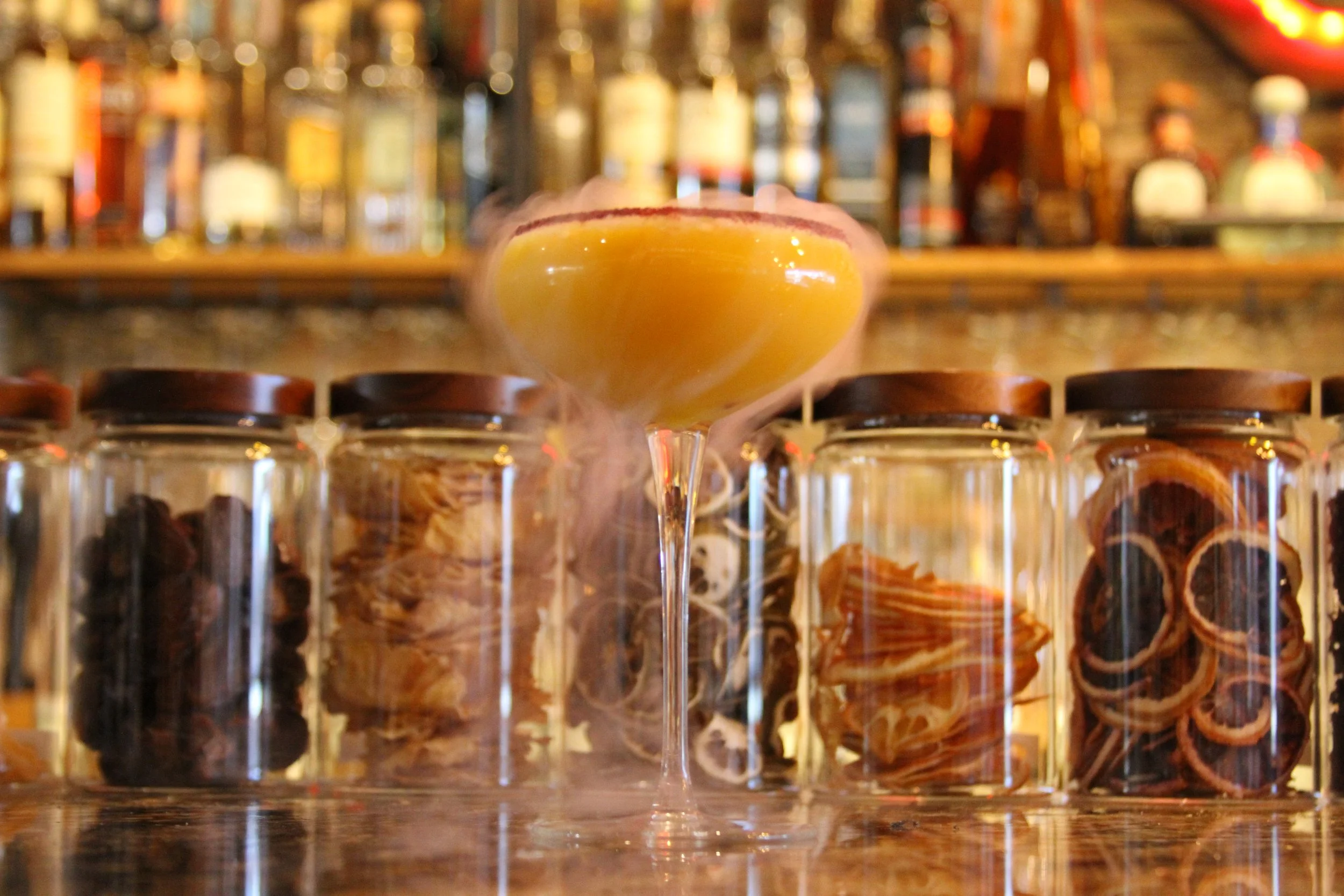 A cocktail glass with a yellow-orange drink, surrounded by jars of ingredients, with blurred bottles in the background.
