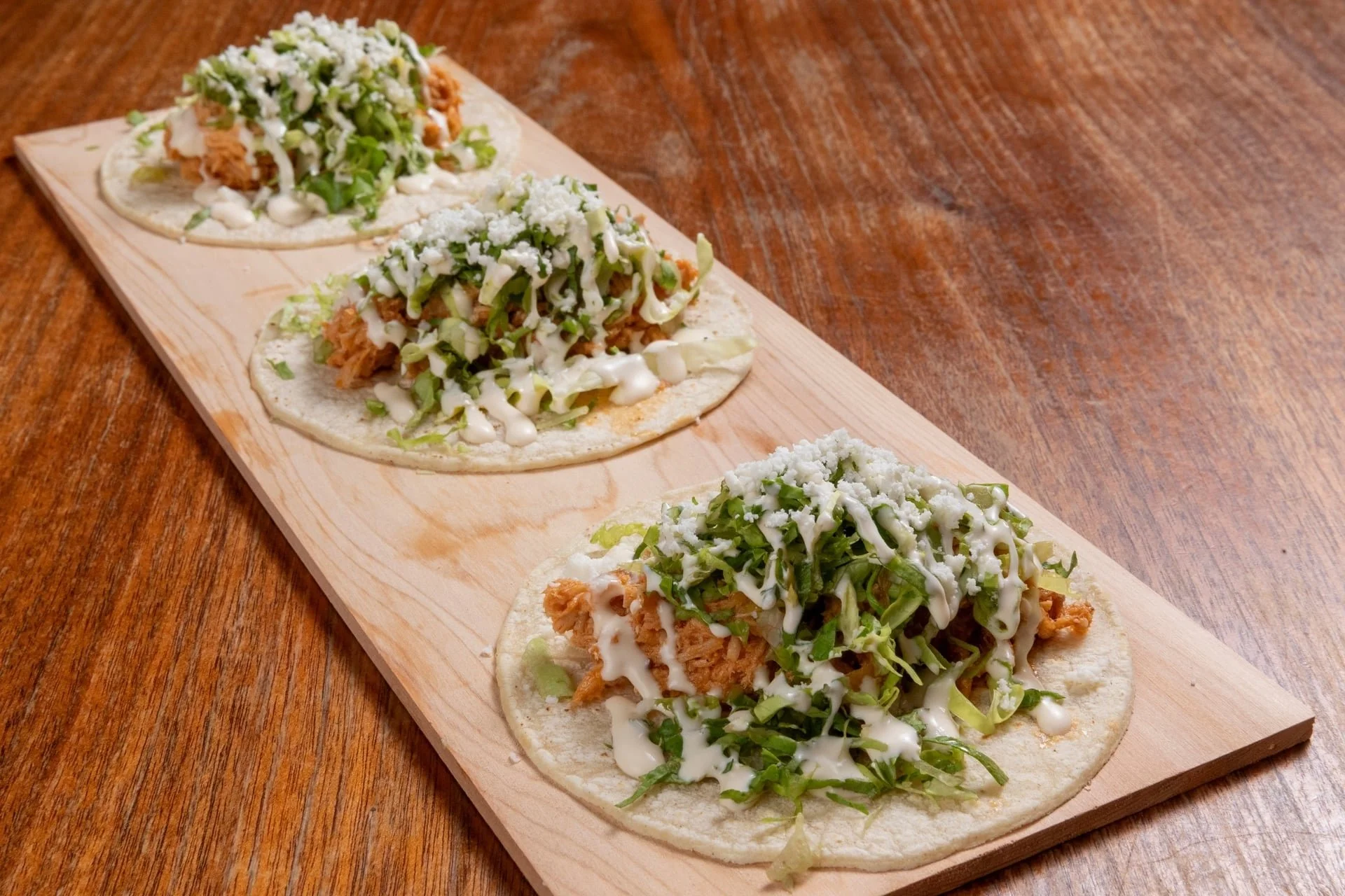 Three fish tacos on a wooden serving board topped with shredded lettuce, shredded cheese, and drizzled with white sauce, on a wooden table.