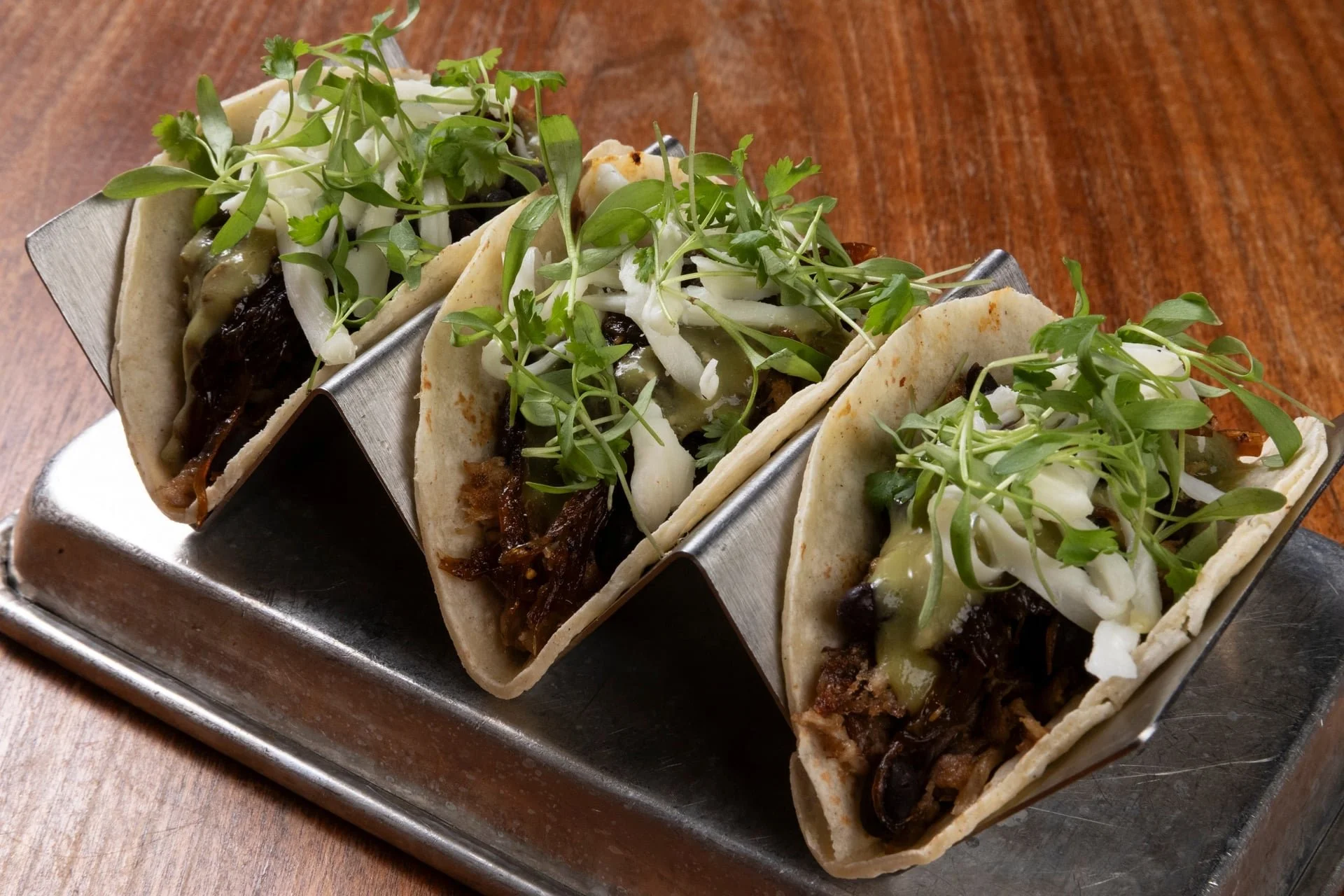 Three tacos filled with shredded meat, cheese, and topped with microgreens on a metal tray, placed on a wooden surface.