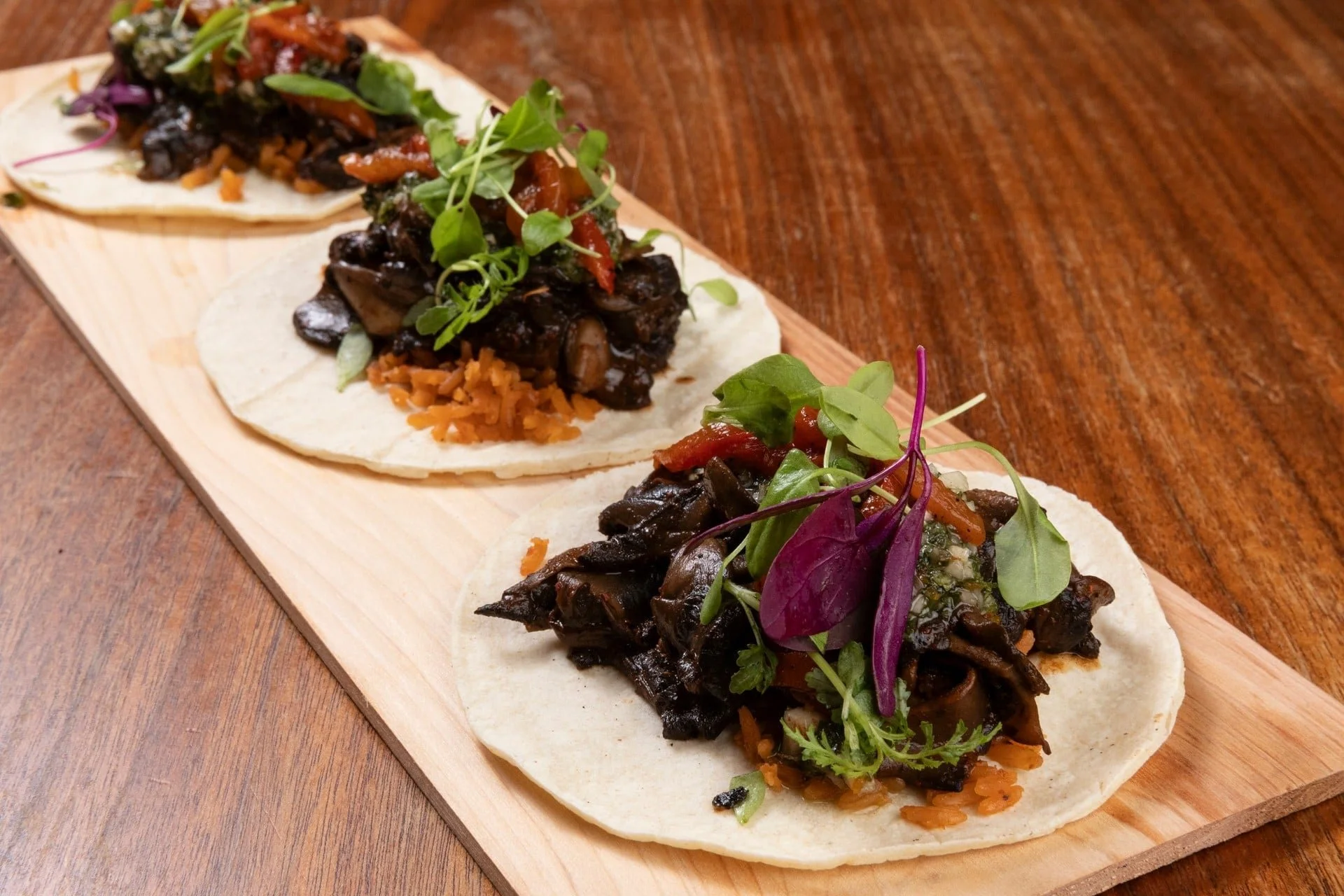 Three small tacos with black beans, vegetables, and microgreens on a wooden serving board.