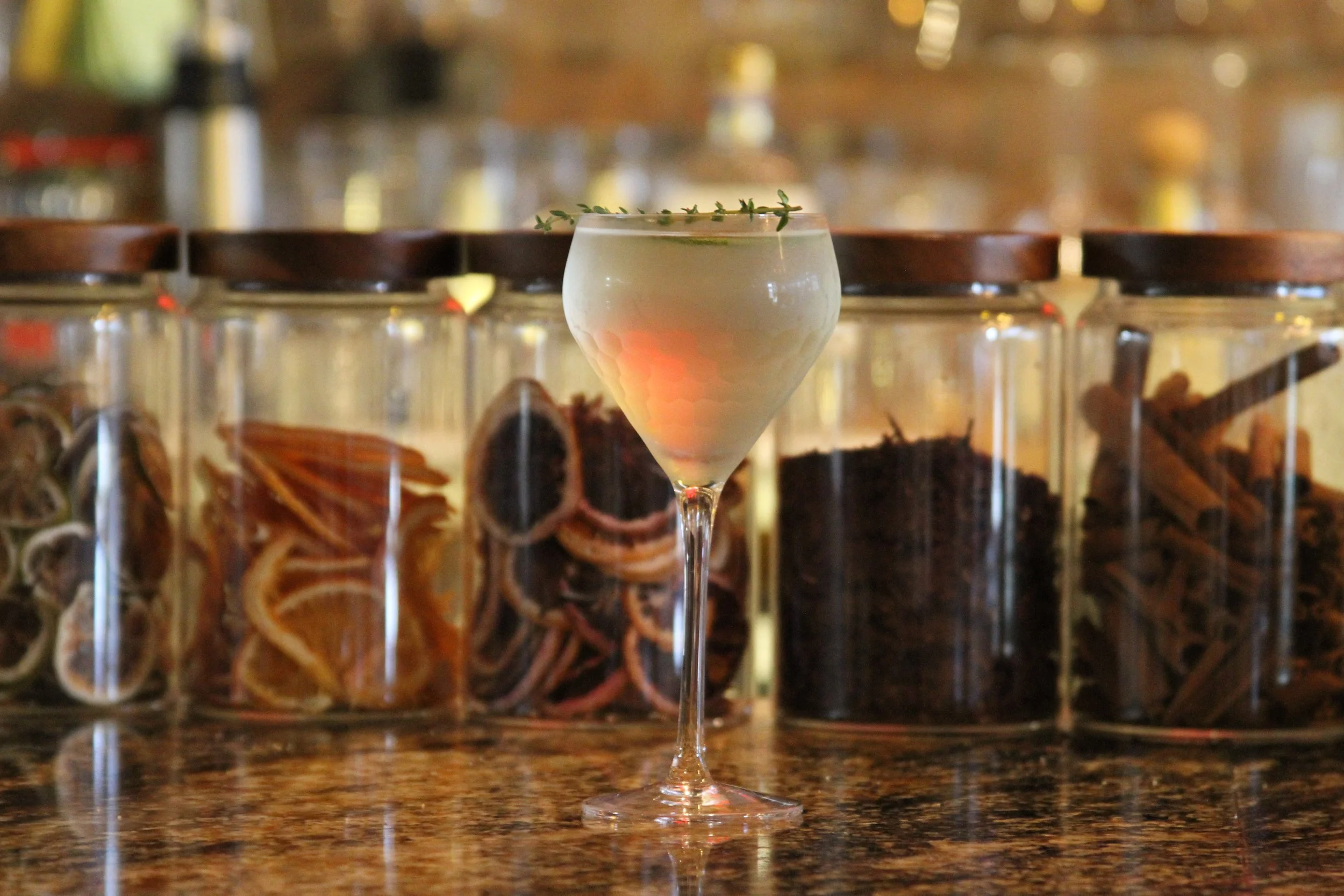 A cocktail with a sprig of herbs served in a martini glass, with jars of dried herbs or spices in the background.