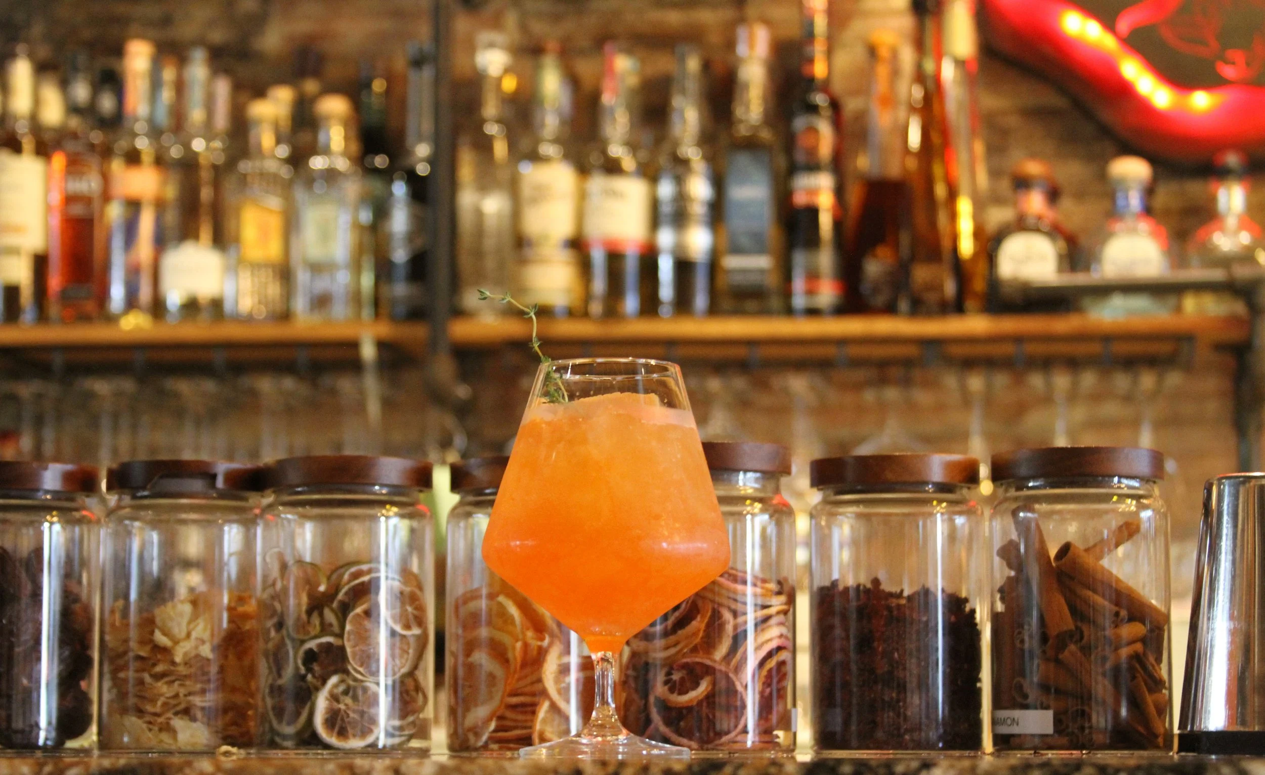 A bar with a background full of liquor bottles and glass jars filled with dried herbs and spices. In the foreground, a cocktail with an orange hue, garnished with a sprig of thyme, is placed on the counter.