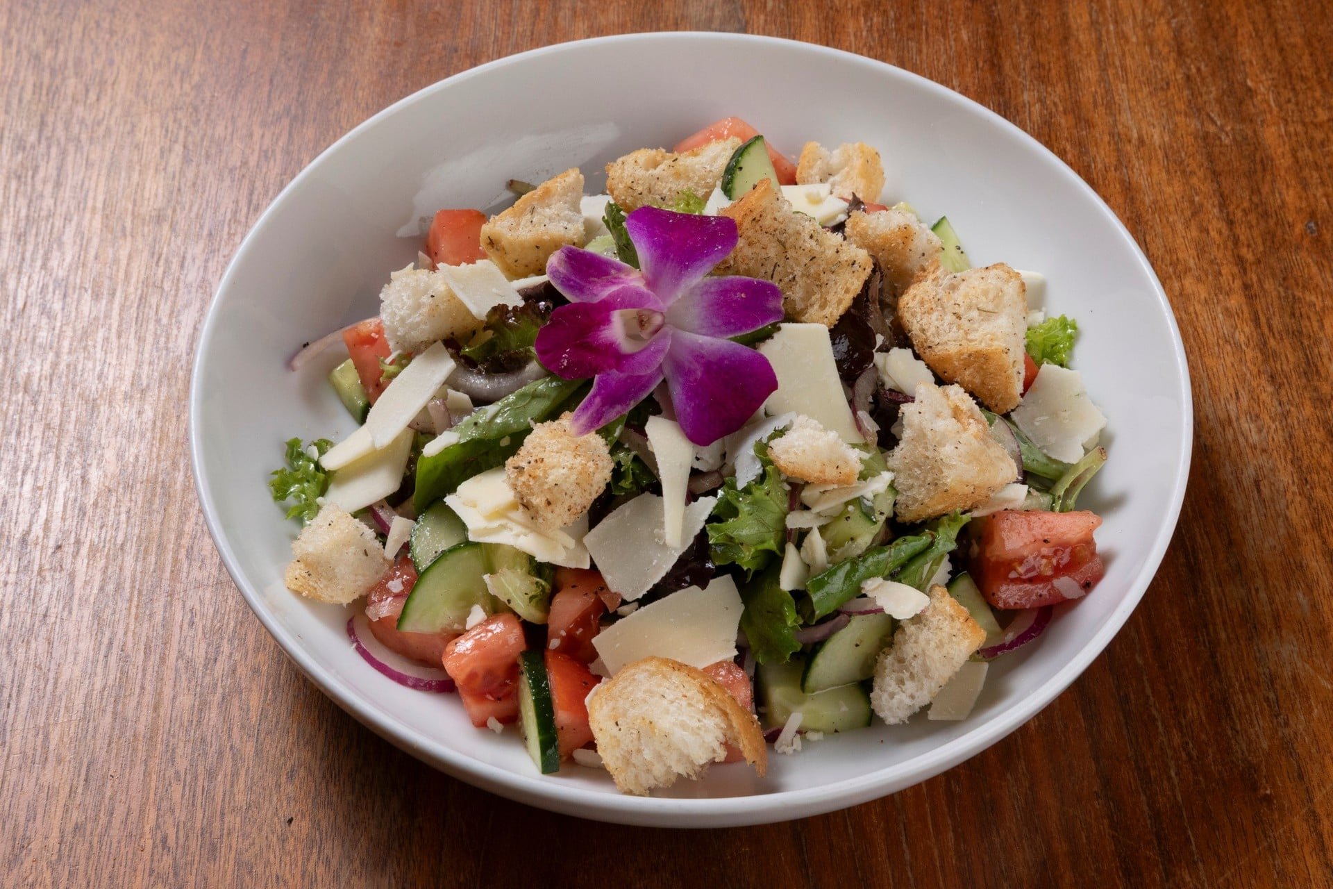 A bowl of fresh salad with lettuce, tomatoes, cucumbers, shredded cheese, croutons, and a decorative purple flower on top, placed on a wooden table.