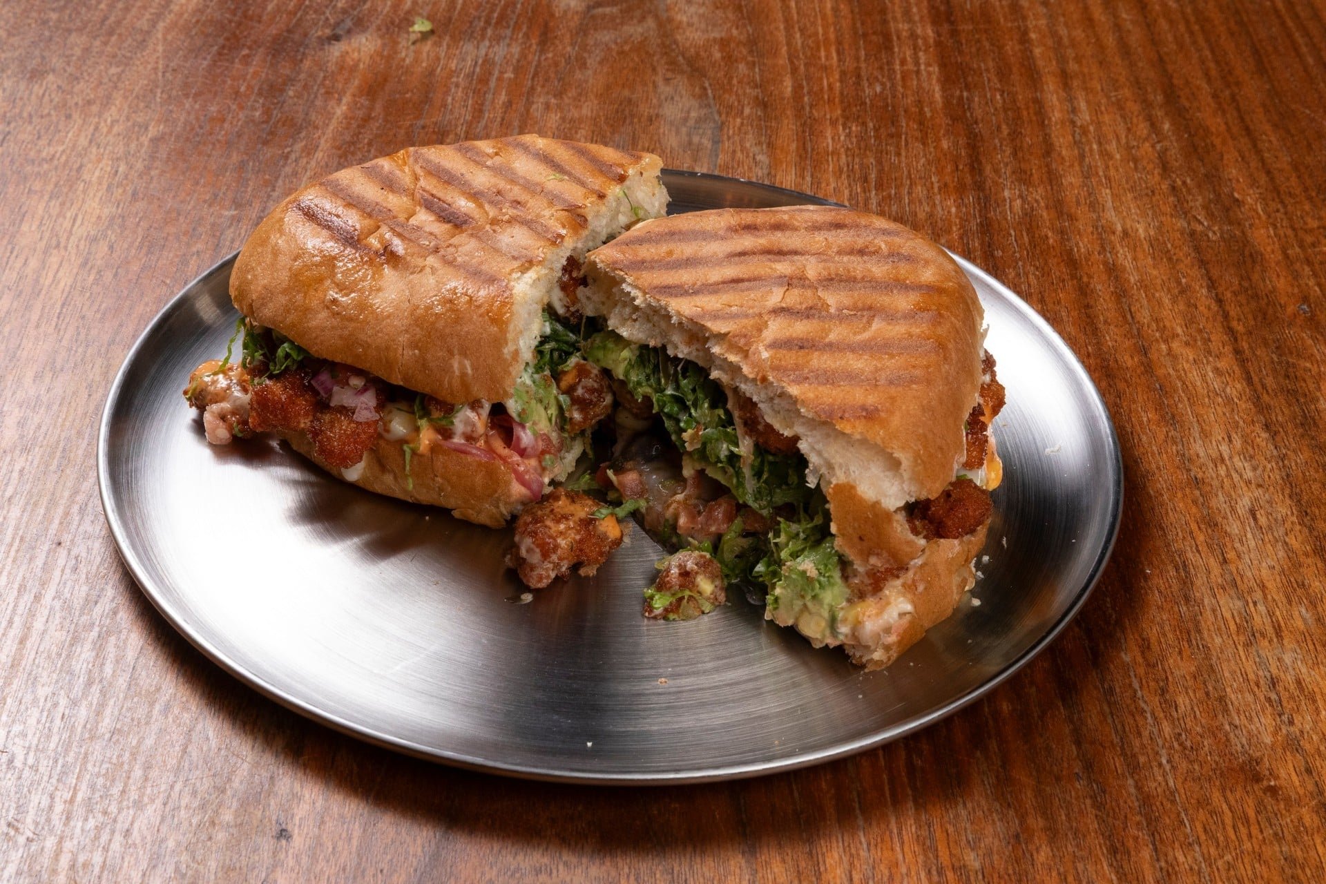 A pressed sandwich on a metal plate with toasted bread and fresh vegetables inside, placed on a wooden table.