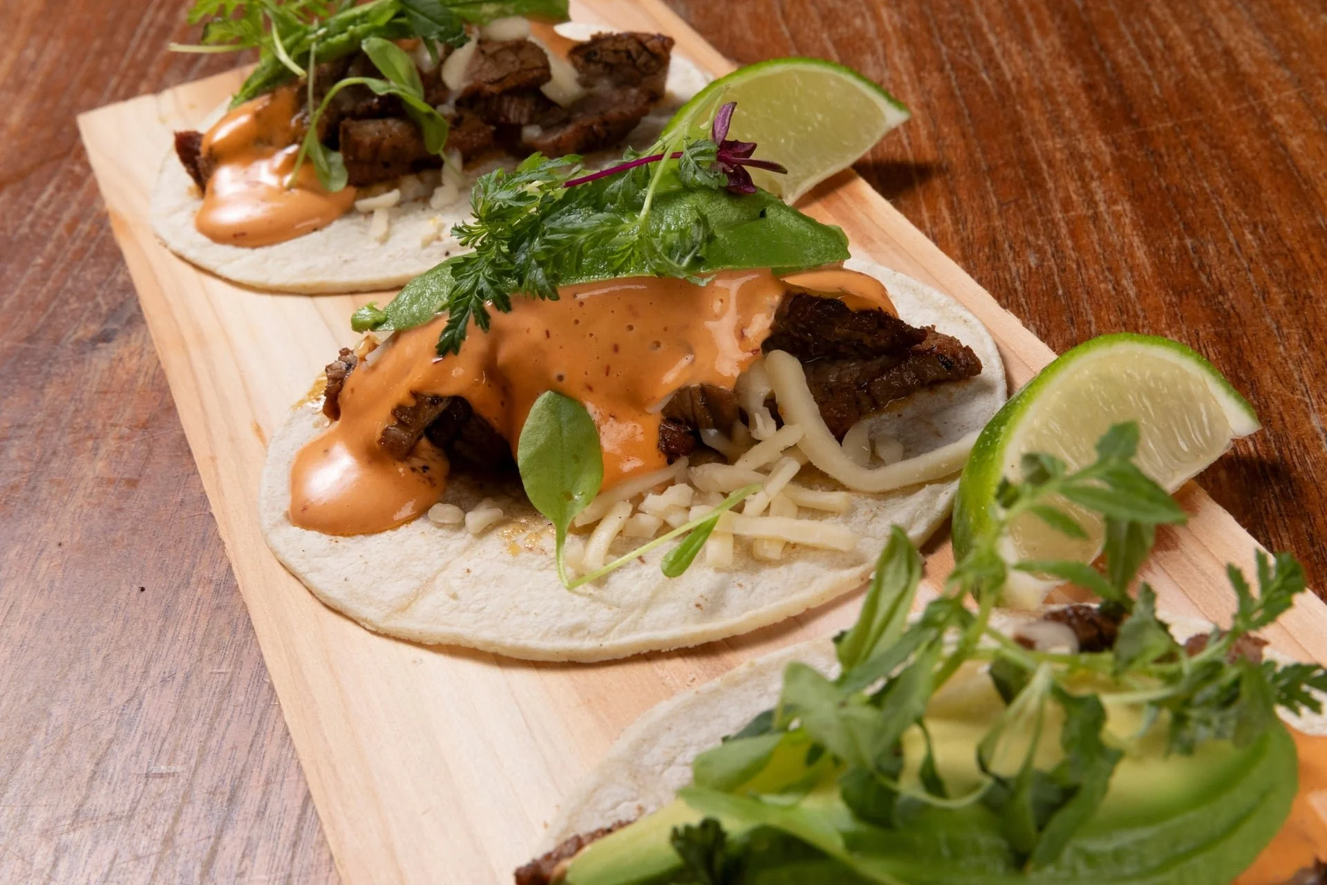 Three street tacos with grilled beef, lettuce, cheese, and creamy sauce, garnished with lime wedges and fresh herbs on a wooden serving platter.