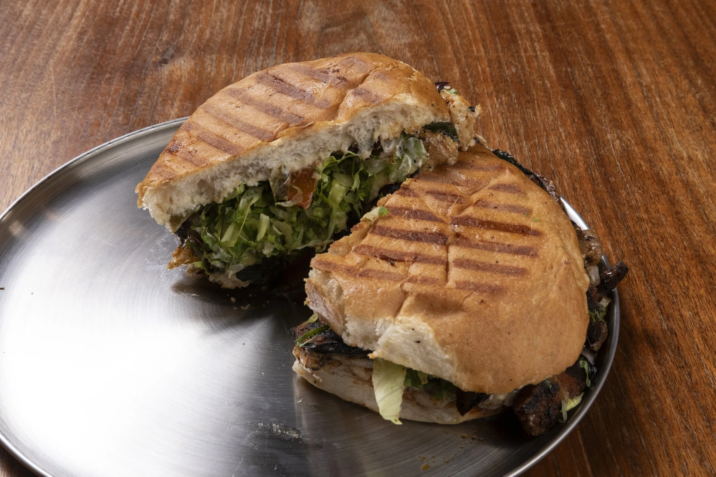 A grilled sandwich with visible lettuce and vegetables, cut in half, placed on a round metal plate on a wooden table.