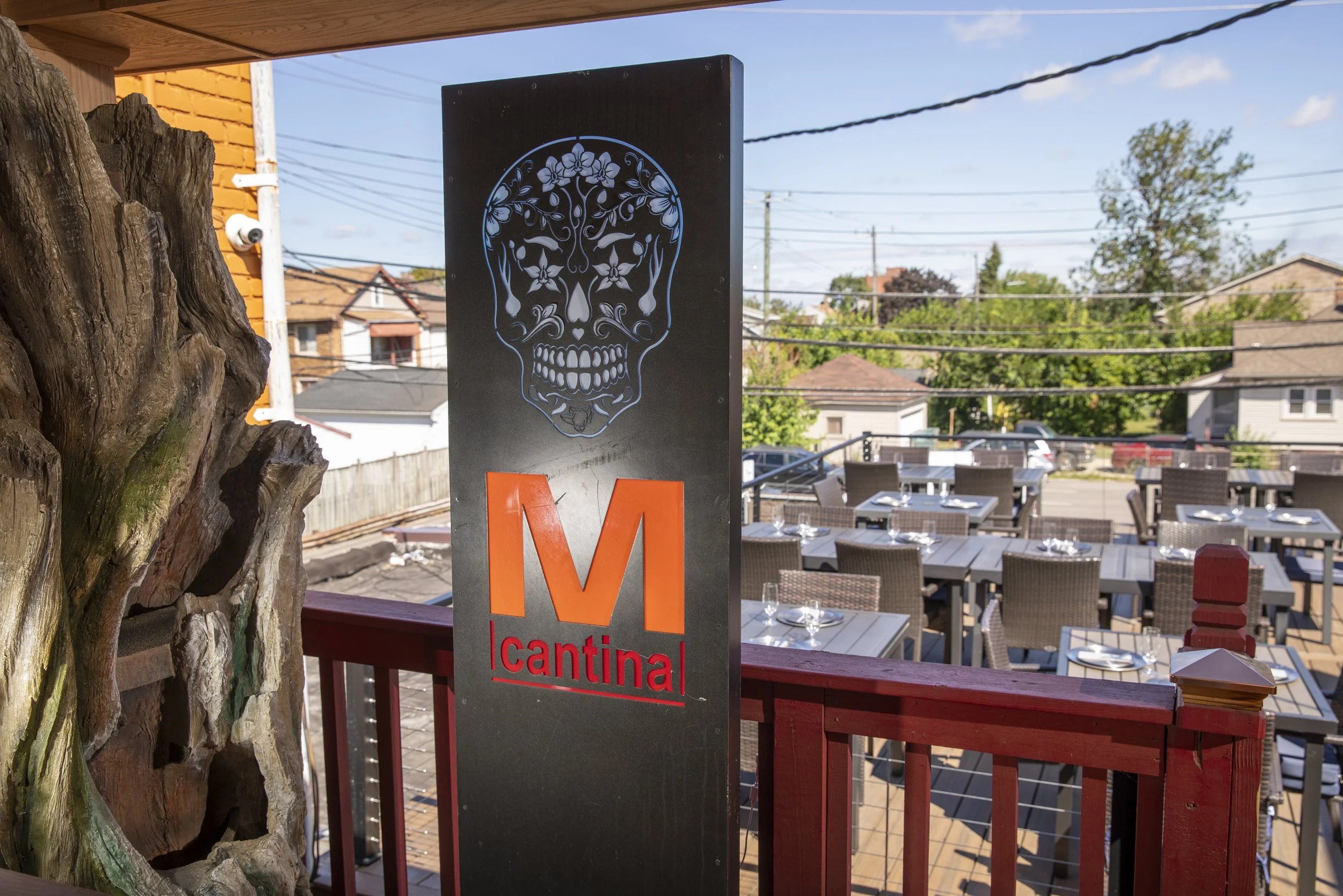 Outdoor patio area with tables set for dining, a sign that reads 'M cantina', and a decorative skull design on a sign. Residential houses and trees are visible in the background.