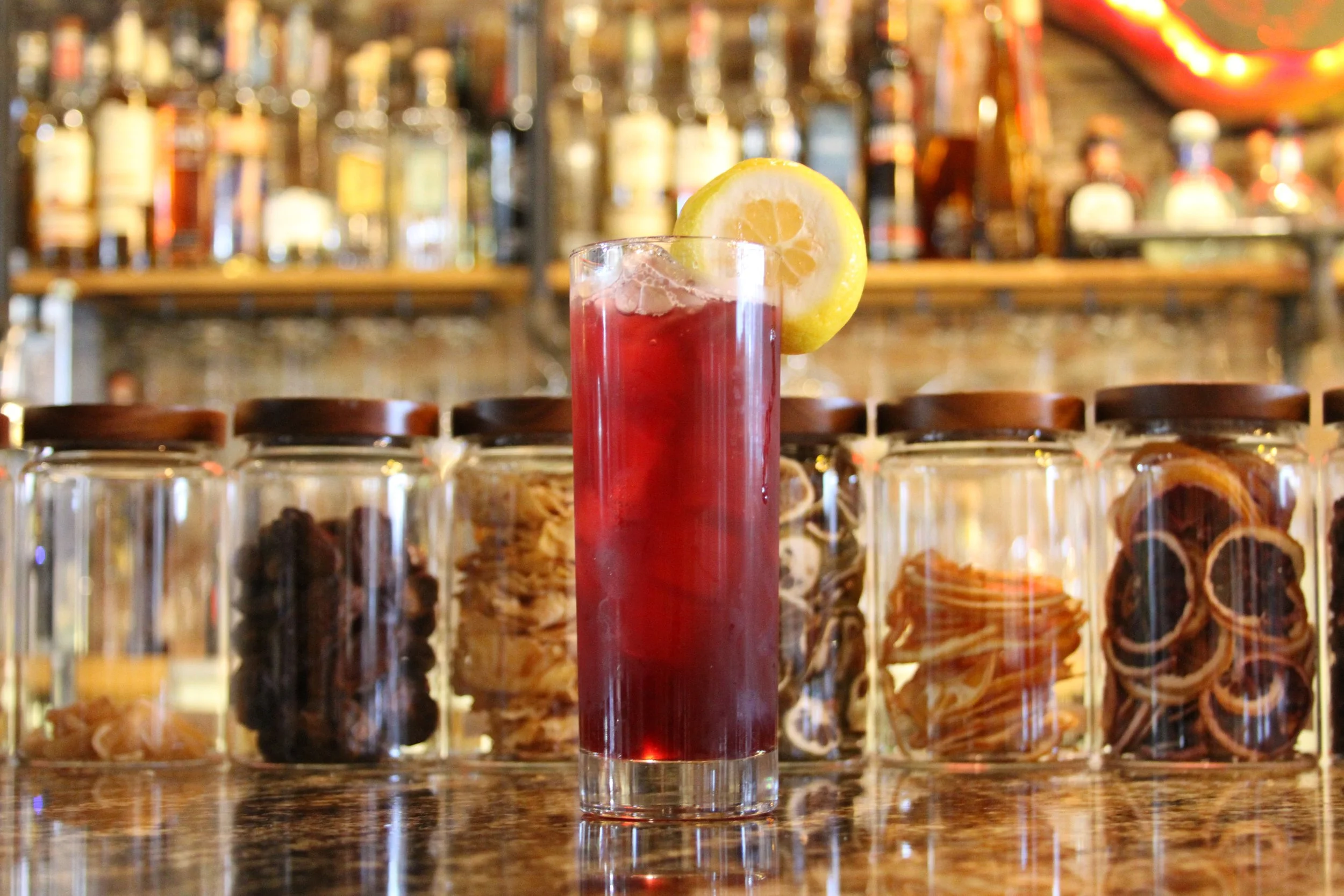 A tall glass with a red beverage and a lemon slice on the rim, in front of a bar shelf with various bottles.