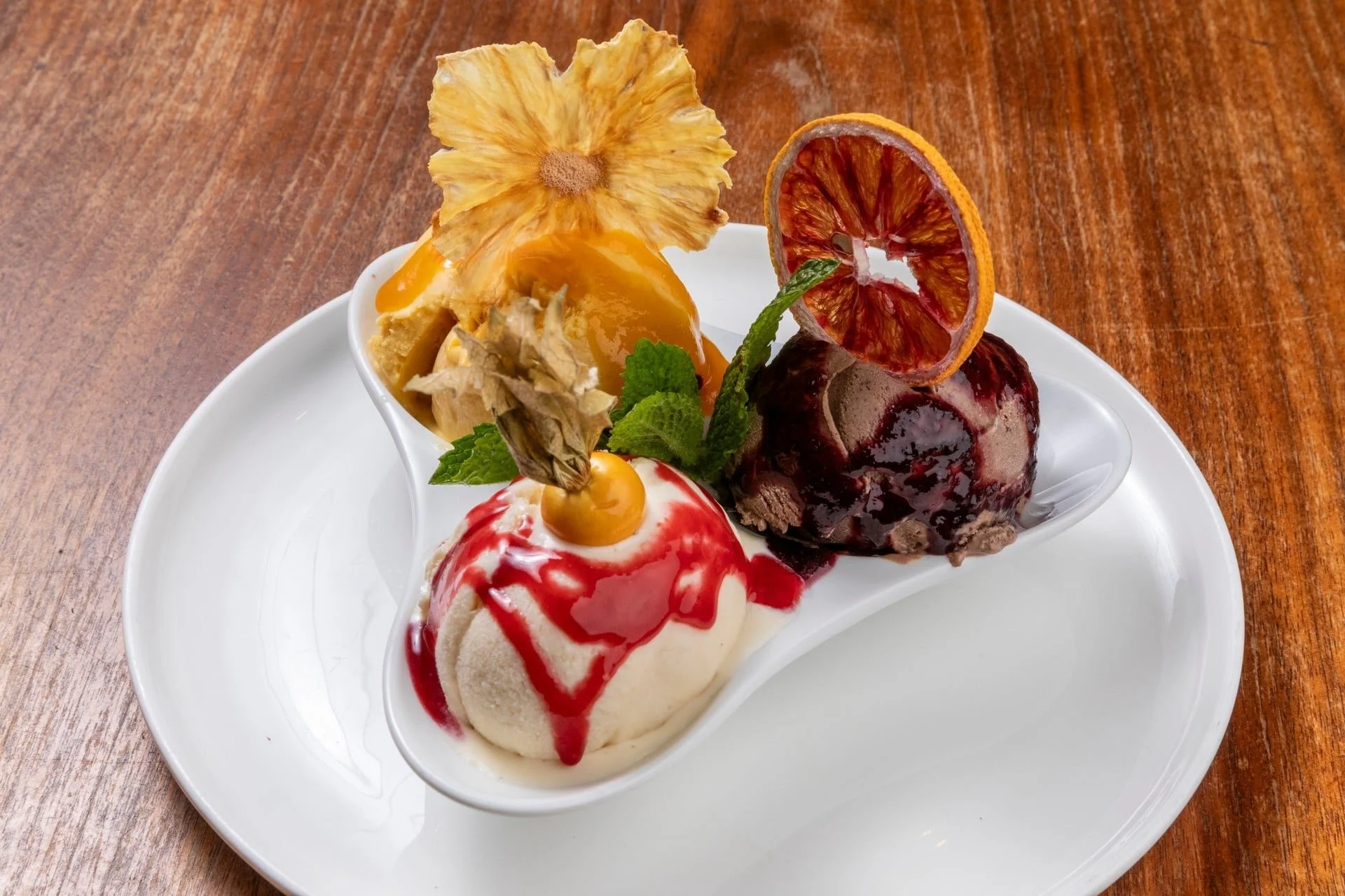 A white oval dish with several pieces of ice cream topped with fruit, syrup, and garnishes, inside a white bowl on a wooden table.