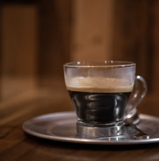 A glass espresso cup filled with black coffee, resting on a saucer with a spoon, on a wooden table with a wooden background.