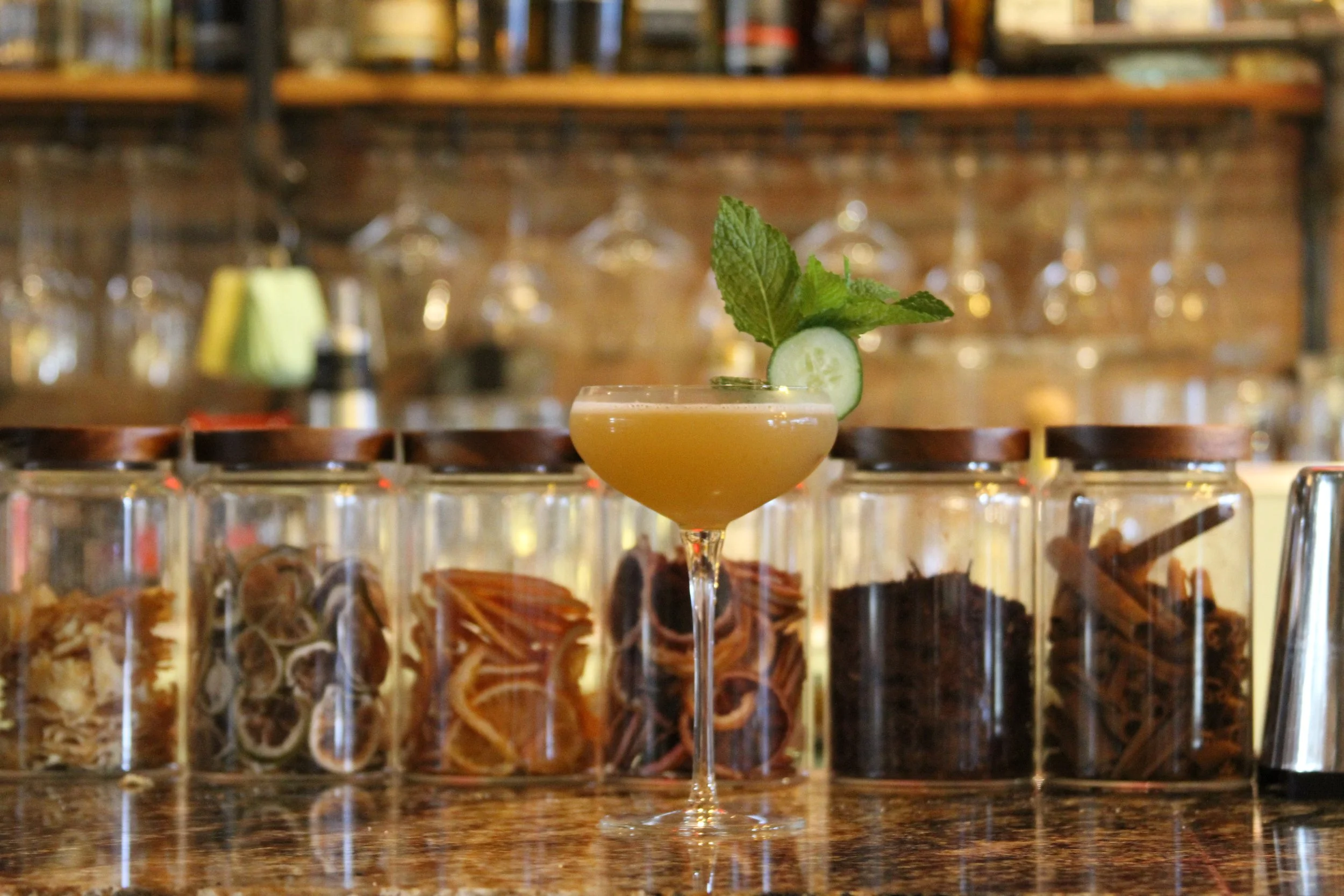 A cocktail in a stemmed glass garnished with a cucumber slice and mint leaves, set on a bar counter with liquor bottles and glassware in the background.
