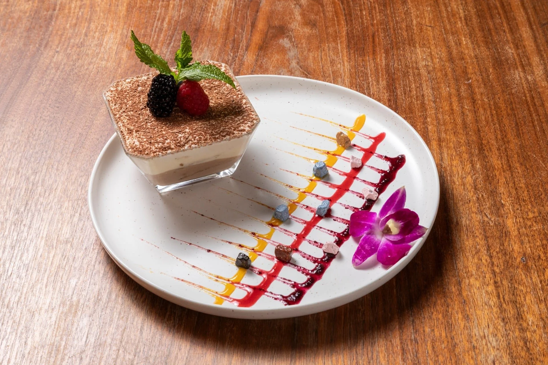 A dessert in a glass cup, topped with blackberries, raspberries, and mint leaves, served on a white plate decorated with colorful sauces, edible flowers, and small candies on a wooden table.