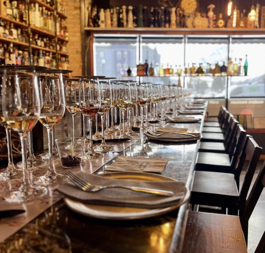 A bar or restaurant interior with a long table set with wine glasses, plates, and cutlery, with shelves of bottles on the wall and large windows overlooking city buildings.