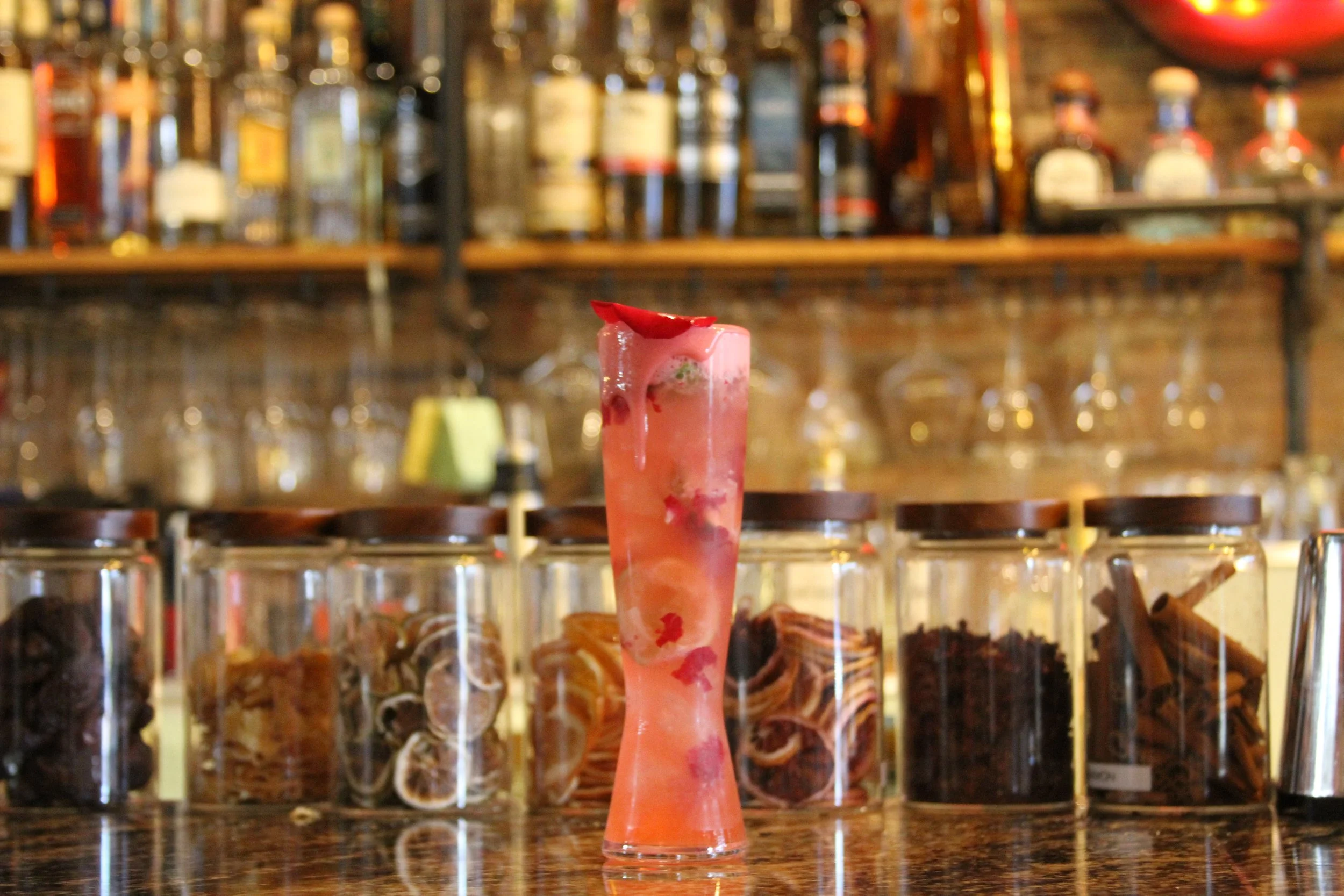 A pink cocktail with a red garnish in a tall glass, set on a bar counter with a background of liquor bottles on shelves.