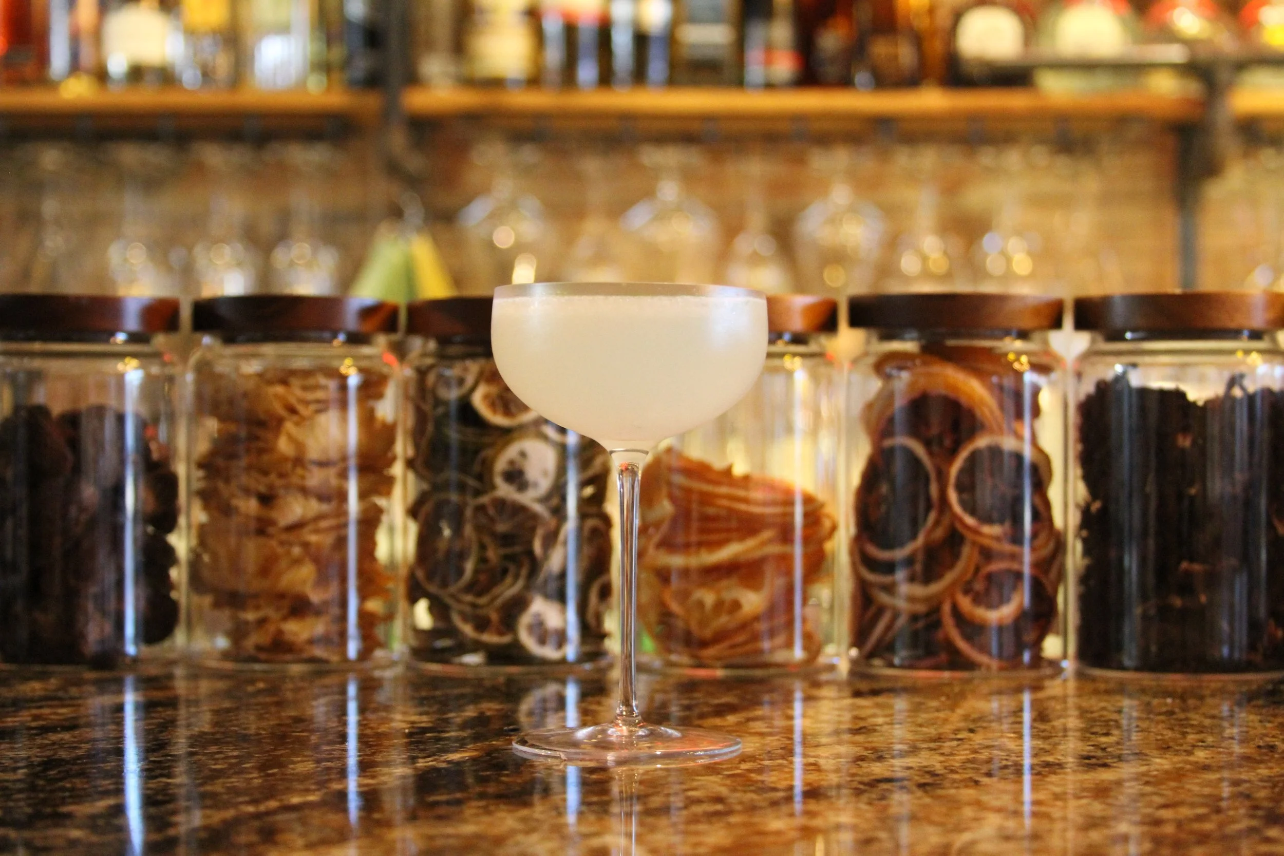 A cocktail glass with a pale yellow drink on a bar counter, with glass jars filled with ingredients and a shelf of liquor bottles in the blurry background.